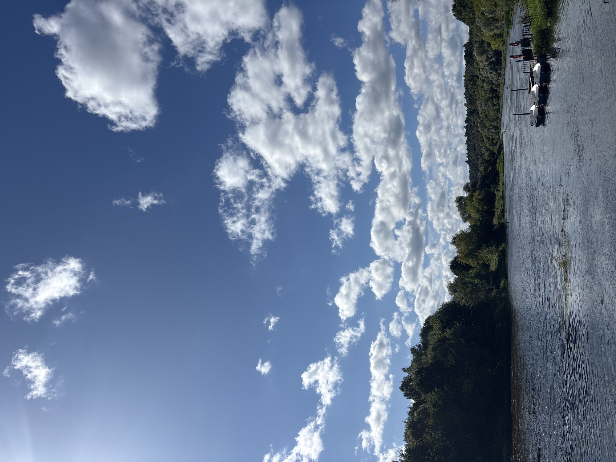 This image captures a serene view of the Moskva River in Zvenigorod, Russia. The scene is dominated by a vast blue sky filled with fluffy white cumulus clouds, suggesting a pleasant day. The river flows calmly, reflecting the sky and surrounding lush green trees that line its banks. On the right side of the river, a small dock is visible, with several white boats moored to it. The boats appear to be small rowboats or kayaks, some with visible numbers. In the distance, the tree-covered hills of Zvenigorod form a picturesque backdrop. The overall atmosphere is tranquil and idyllic, showcasing the natural beauty of the location.