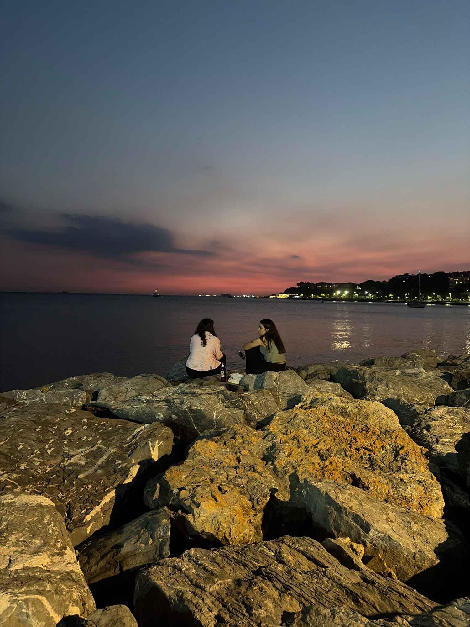The image captures a twilight scene on a rocky shoreline in Kadıköy, Turkey. Two young women are seated on large, weathered rocks overlooking the calm sea. The sky displays a gradient of deep blue to a soft pink and orange as the sun sets, with scattered clouds. In the distance, lights from the cityscape twinkle along the horizon, and a boat is visible on the water. The foreground is dominated by the texture and varied colors of the rocks, some appearing damp and dark, others lighter and tinged with orange. One woman wears a light-colored, patterned shirt and dark pants, while the other is dressed in a light green top and dark pants. A bottle and a small white object are placed on the rock between them. The overall atmosphere is serene and peaceful, suggesting a moment of quiet contemplation or conversation.