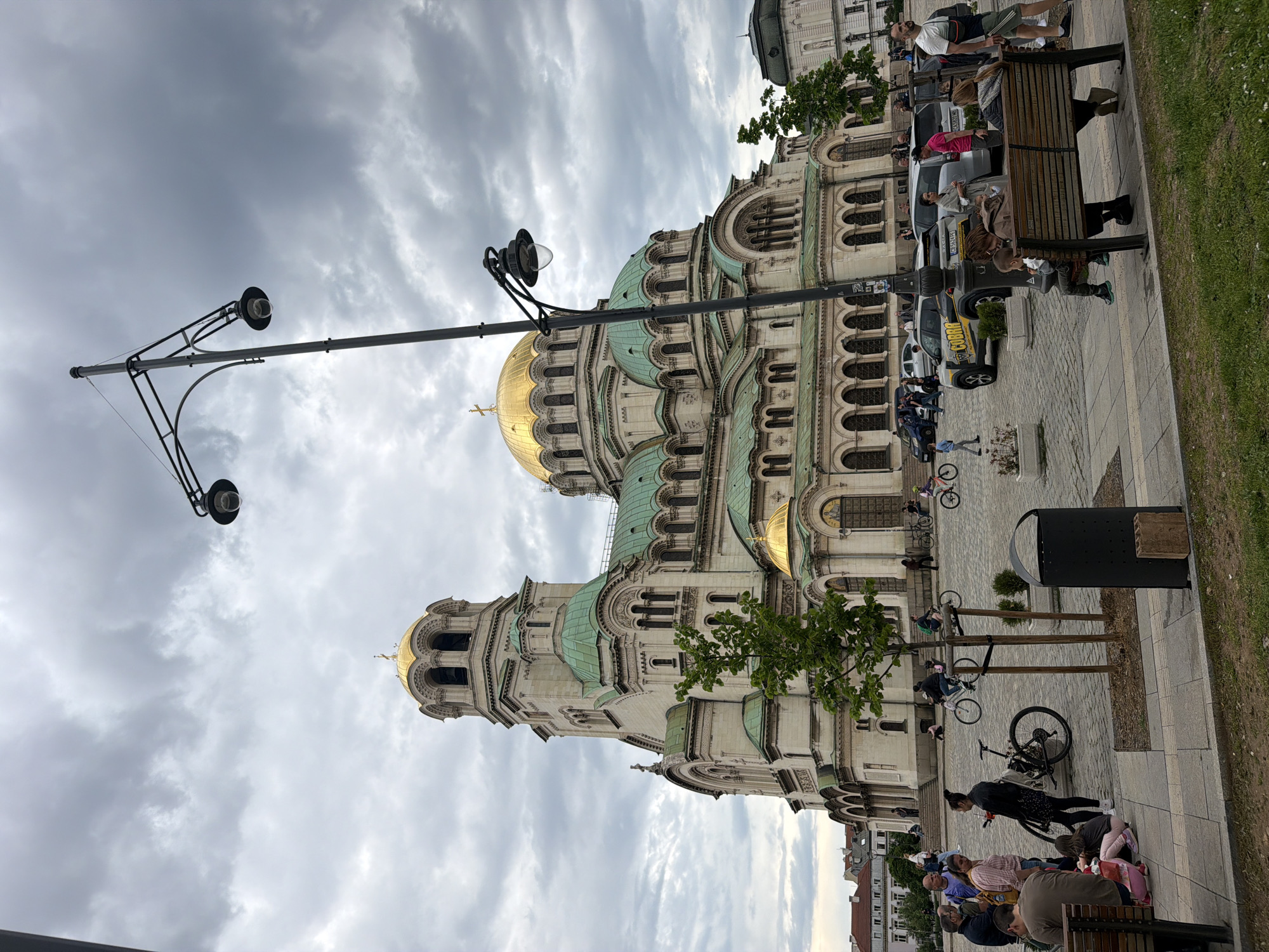 The image captures a street scene in Sofia, Bulgaria, dominated by the Alexander Nevsky Cathedral. The cathedral, with its gold domes and green roofs, stands prominently against a cloudy sky. The foreground features a paved area with benches where people are sitting and relaxing. Some are on bicycles. Street lamps are also visible. Vehicles are seen further back near the building.