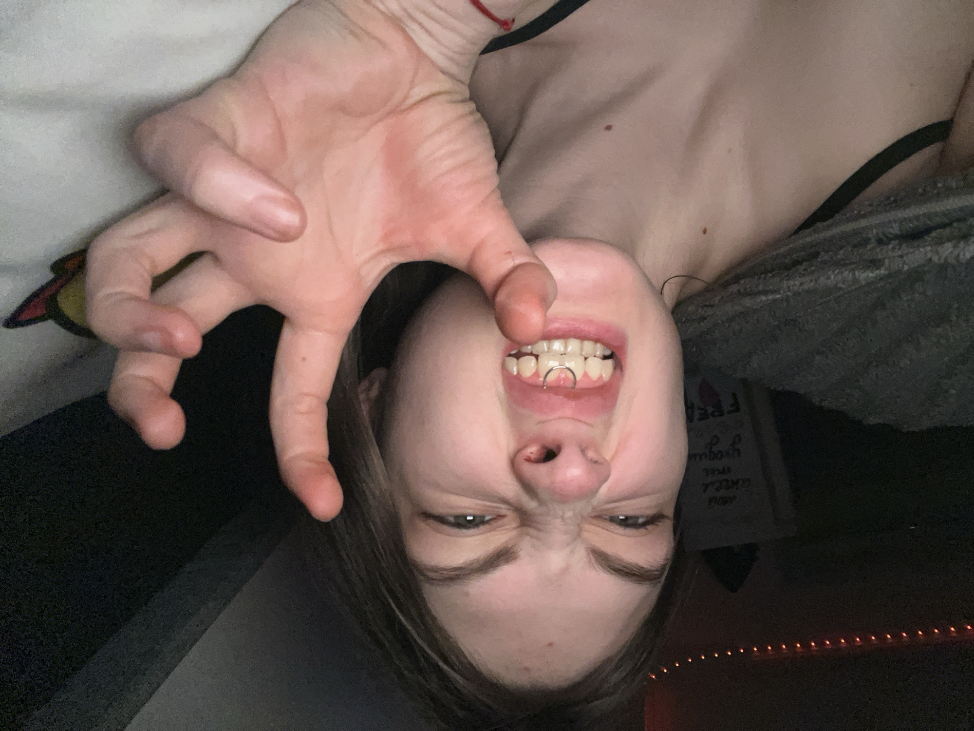 A person is upside down, positioned at the top of the frame, grimacing with their hand extended toward the camera in a claw-like gesture. They have light skin and appear to have a piercing between their front teeth. A black tank top is visible. The background shows a partial view of a room with indistinct objects, including a sign and string lights. No information connects this image to Saint Petersburg, Russia.