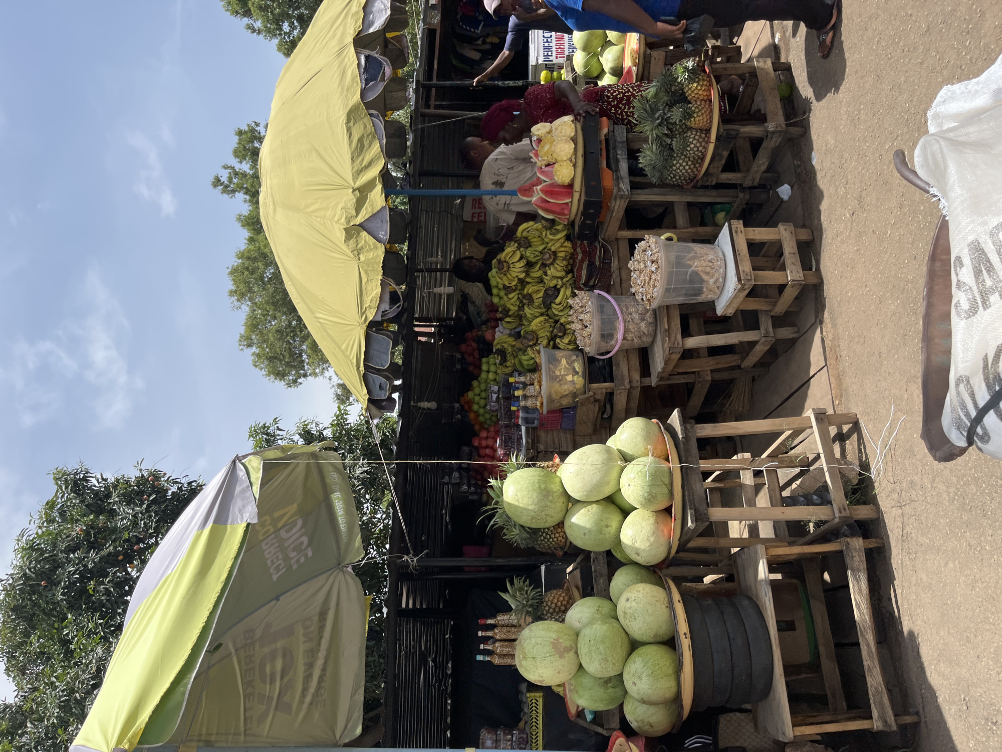 The image depicts a roadside fruit stand in Jos, Nigeria. The stand is stocked with a variety of fruits including watermelons, pineapples, bananas, and coconuts, displayed on wooden crates. Vendors are visible behind the fruit displays. The stand is covered by a yellow umbrella providing shade. Additional trees and umbrellas can be seen in the background.