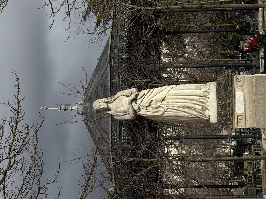 The image depicts a white stone statue of Sainte Genevieve standing centrally in the Jardin du Luxembourg, Paris, France. The statue, appearing worn with age, shows a female figure with long braided hair, draped clothing, and hands clasped at her waist, looking downwards. The inscription "SAINTE GENEVIEVE PATRONNE DE PARIS NÉE 423 MORTE 512" is visible on the statue's stone pedestal.
Behind the statue, an ornate hexagonal bandstand (kiosque à musique) with a dark, sloped roof and decorative dark metal railings dominates the mid-ground. A white seagull is perched atop the bandstand's finial. The surrounding environment consists of numerous leafless deciduous trees, indicating a late autumn or winter season.
The sky is overcast with dark, dramatic grey clouds. In the background, partially obscured by the trees and bandstand, are light-colored buildings. Several individuals are present in the lower frame: a person is seated on the left near a stack of green outdoor chairs, and another individual wearing a red jacket is seated on the right, also at an outdoor table. Black lampposts are visible among the trees.