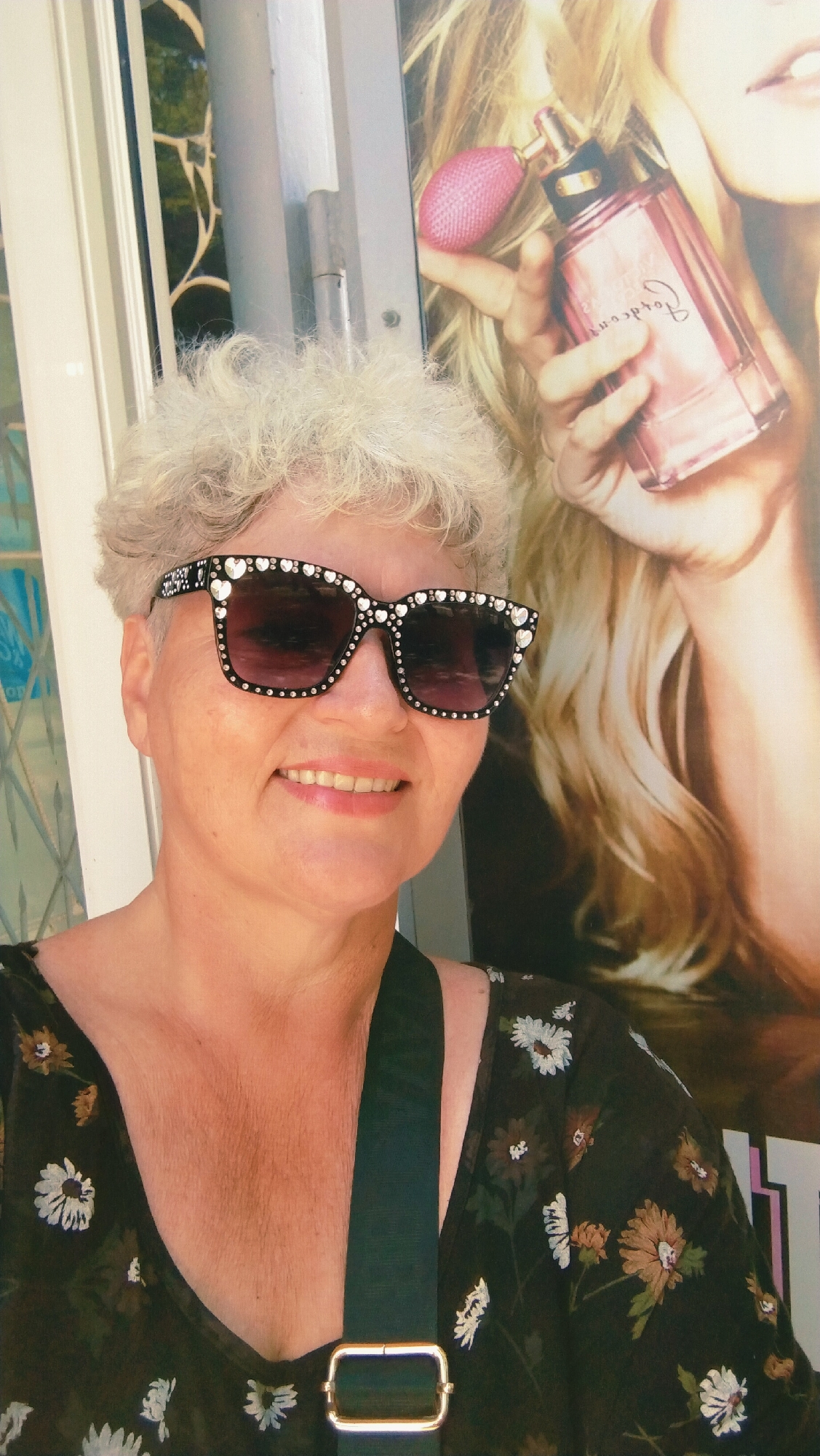 A woman is standing in front of a large advertisement for perfume, holding her phone and taking a selfie. She has short, curly, blonde hair, and is wearing large, embellished sunglasses with rhinestones and heart-shaped gems. She is also wearing a dark, floral-patterned top. A black strap with a gold buckle is visible across her chest, part of a bag she is carrying. The background features the perfume advertisement, which depicts a woman with blonde hair holding a pink perfume bottle. The location is identified as the city of Horishni Plavni, Ukraine, suggesting this scene is likely in a public area within the city, possibly a shopping center or street with advertisements. The lighting is bright and appears to be natural sunlight.