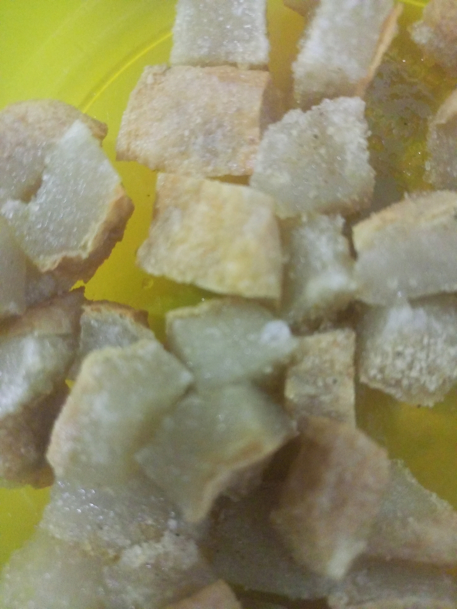 The image is a close-up, top-down view of a pile of fried pork belly pieces in a bright yellow bowl. The pork belly pieces are irregular in shape and size, and appear to be coated in a light, crispy batter. Some pieces show visible layers of fat and meat. The lighting is bright, causing some glare on the oily surface of the food. There are no people or any other objects visible in the frame. The scene is likely indoors, given the artificial lighting. The location in Poka'an, Indonesia, is not depicted in the image itself.