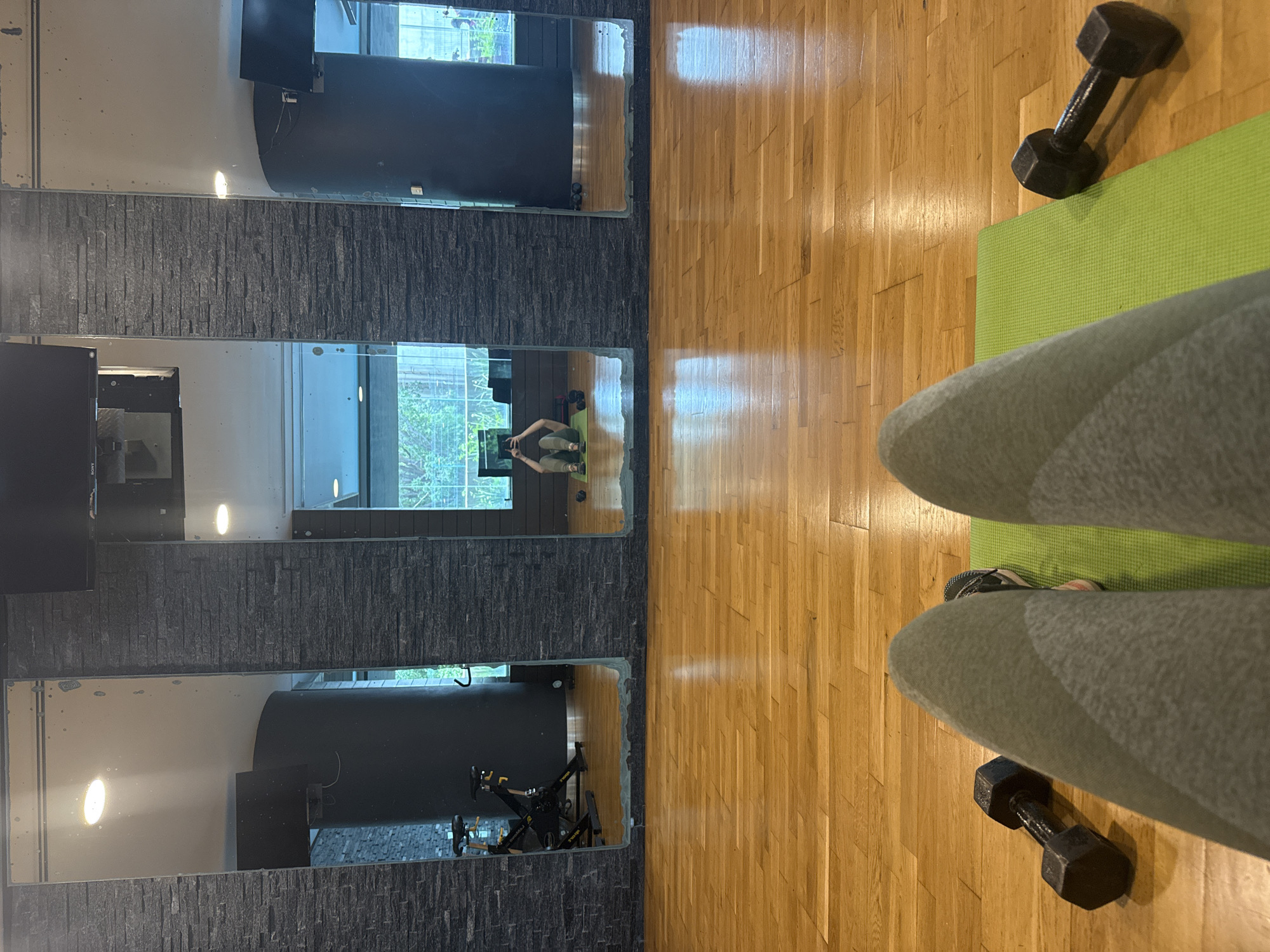 The image shows a person standing on a green yoga mat with dumbbells on either side, likely indoors in a gym or fitness area. The floor is made of hardwood. The person is wearing grey sweatpants and sneakers, partially visible. Behind the person and yoga mat are three vertical mirrors framed by a wall with dark grey brick or stone-like texture. The mirrors reflect the room's interior, showing a window with an outdoor view of trees and what appears to be another person sitting or exercising in the mirror reflection. Another mirror reflection includes the equipment and interior of the fitness area. Based on the view of the window, it is difficult to assess the location of the scene in Mexico City, Mexico, because there is not enough information about the external view.
