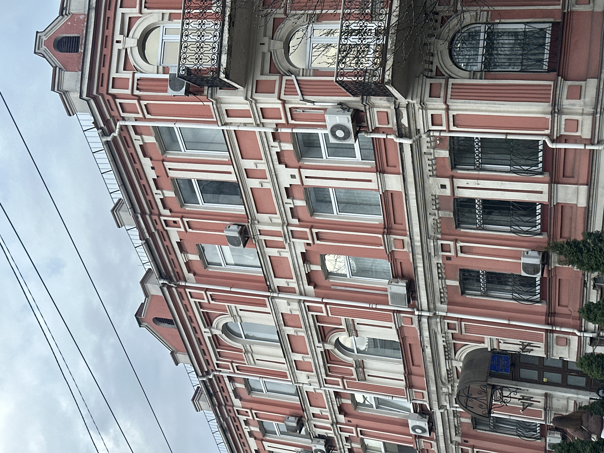 Here is a description of the image:
The image shows a building in Kyiv, Ukraine. It is a multi-story building with a pink and white facade. The building has windows of various shapes and sizes. There are air conditioning units installed on the exterior walls of the building. There are power lines visible in the sky. The sky is overcast.
