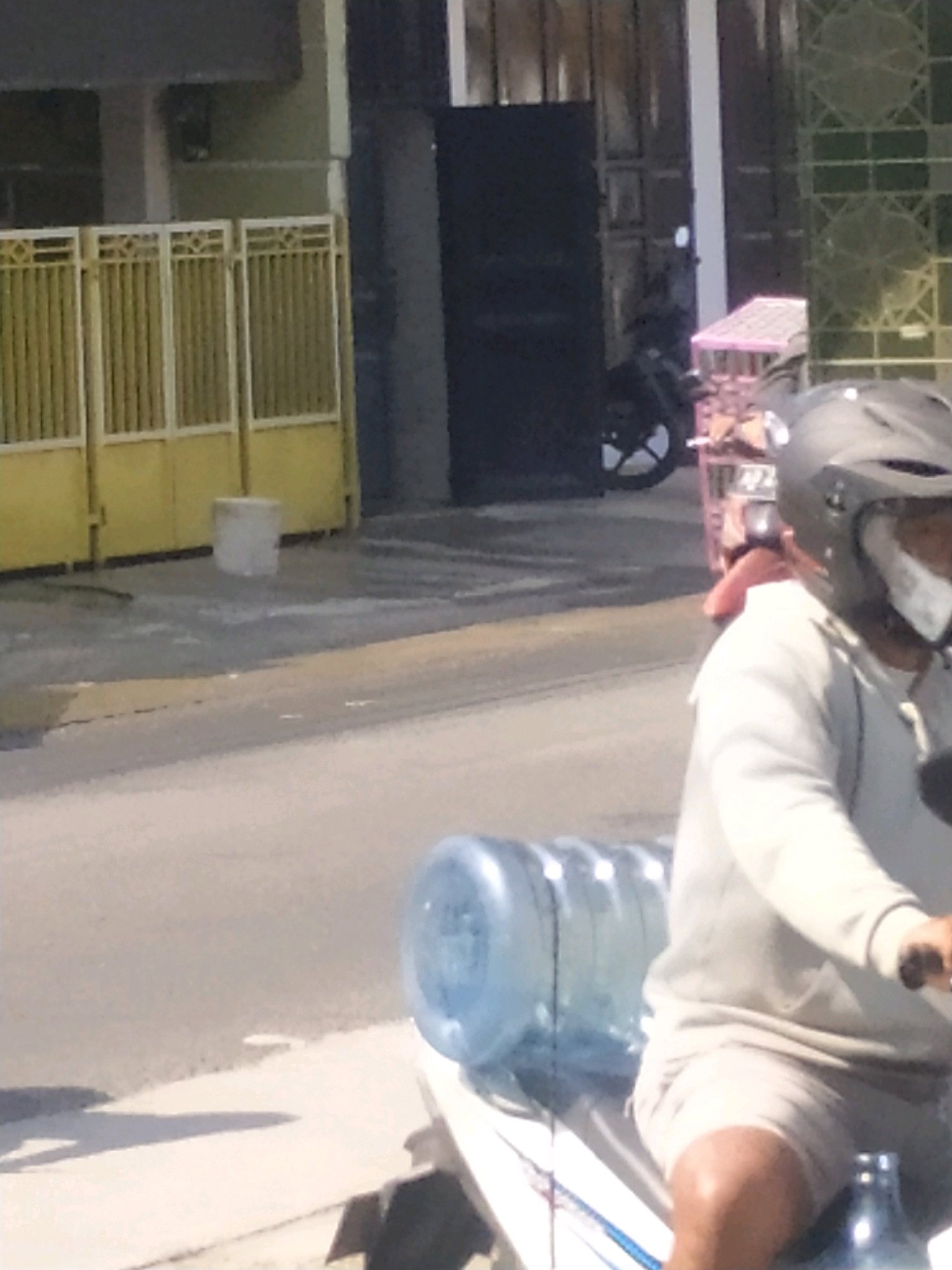 A person wearing a black helmet and a light-colored long-sleeved shirt is riding a motorcycle. The motorcycle is carrying a large blue water jug and another smaller jug is visible. The background includes a yellow gate, a concrete block, and a building. The scene is outdoors, on a paved surface that appears to be a street or pathway. The image is grainy and out of focus, making detailed identification difficult.