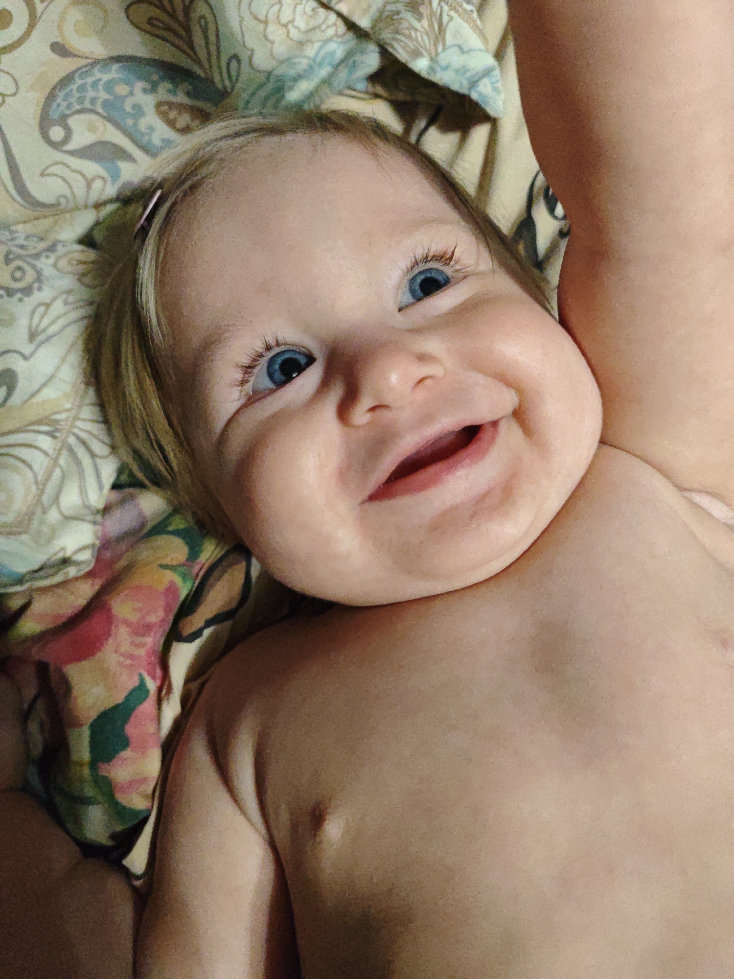 The image shows a close-up, overhead view of a baby's face and upper torso. The baby has fair skin, blonde hair, and striking blue eyes. The baby is looking upwards and appears to be smiling broadly, with its mouth open. The baby is lying on a patterned surface, possibly a blanket or bedding, with floral and paisley designs in muted colors. To the right of the baby's head, a portion of an adult's arm or leg is visible, also appearing to be fair-skinned. The lighting is soft and natural. There are no discernible landmarks or elements in the image that can be used to determine a specific location in Voronezh, Russia, or any other city.