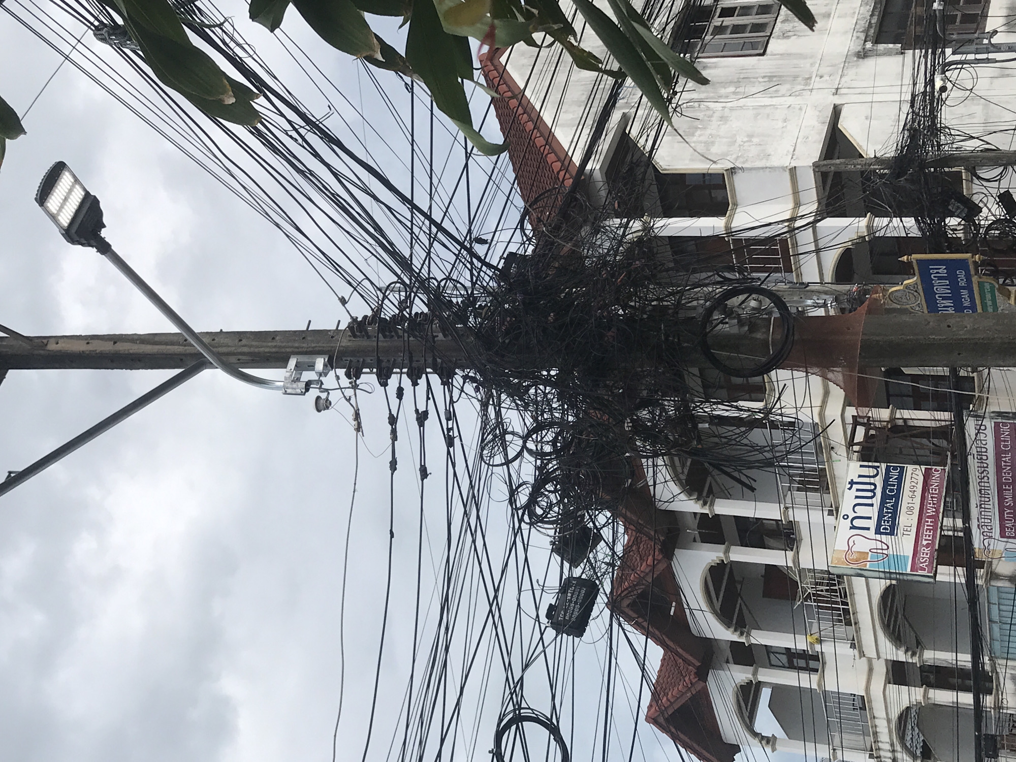 The image depicts a complex tangle of electrical wires and cables suspended from a utility pole in Baan Chaweng, Thailand. The wires are densely packed, creating a chaotic web. A modern LED street light is attached to the pole on a long arm. In the background, a multi-story building with arched balconies and a red-tiled roof is visible. Several signs are attached to the building, including one for "ท่าฟัน DENTAL CLINIC" with a phone number (081-6492779) and the words "LASER TEETH WHITENING." Another sign reads "คลินิกทันตกรรมยิ้มสวย BEAUTY SMILE DENTAL CLINIC." A street sign on the pole reads "มหาดงาม D NGAM ROAD." A small black rectangular object with text "ROS65-00001 20005-00002" is hanging from the wires. Green foliage is visible at the top of the frame. The sky is overcast.