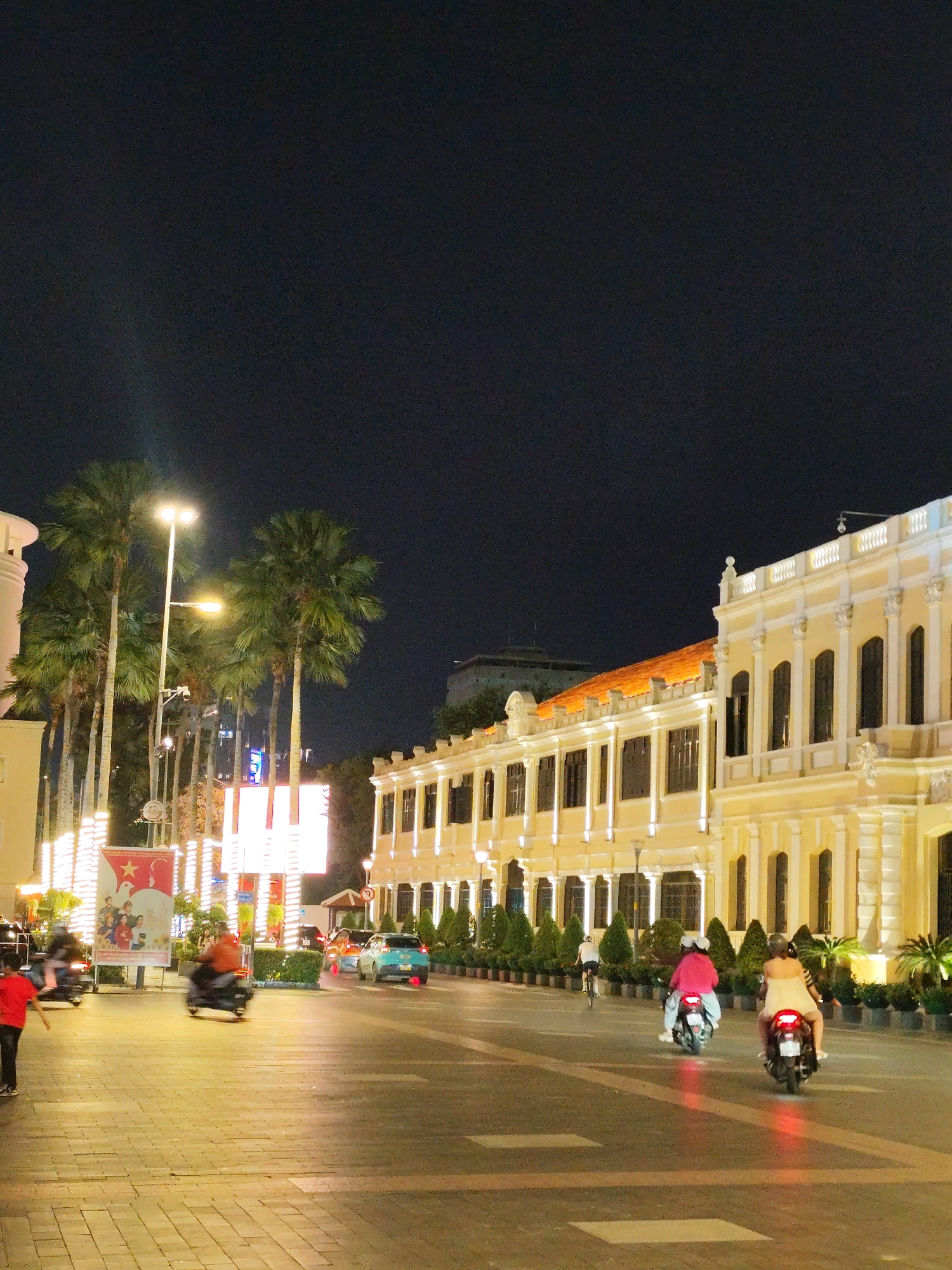 This is a nighttime photograph of a street scene in Ho Chi Minh City, Vietnam, likely taken on a pedestrianized area or a wide boulevard. The image captures a vibrant urban environment illuminated by streetlights and the warm glow of building lights.
In the foreground, a paved walkway with a brick pattern is visible. To the left, a young person in a red shirt and dark pants is running away from the camera. Further down the walkway, several people are riding motorcycles. One motorcycle, ridden by a person in an orange jacket, is moving away from the camera. Another motorcycle, carrying two people, one wearing a pink jacket, is also heading away from the camera. On the right side of the street, a motorcycle with two occupants, the passenger wearing a light-colored dress, is also moving away from the camera, its brake light leaving a red reflection on the road.
The street itself features a few cars, including a light blue vehicle and an orange vehicle, also moving away from the camera. On the left side of the street, a prominent billboard or large poster with a red star and images of people is displayed. Several palm trees are visible along the street, with their fronds illuminated by streetlights.
The background is dominated by a long, yellow building with ornate white detailing, featuring numerous windows and arched doorways. The architecture suggests a colonial-era building, common in Ho Chi Minh City. Another building with a dark, tiled roof is partially visible behind this main structure. The sky is dark, indicating it is nighttime. The overall atmosphere is lively and bustling, characteristic of a city street at night.