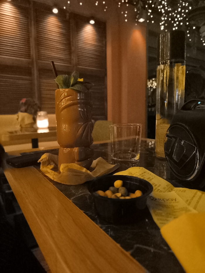 The image presents a close-up, dimly lit scene on a table within an indoor bar or restaurant in Palaio Faliro, Greece, likely during the evening. The central subject is a distinct brown and orange tiki mug cocktail, garnished with fresh mint leaves and a small yellow flower, with a dark straw protruding. It sits on a crumpled yellow napkin on a dark, marble-patterned table.
To the right of the cocktail, an empty, clear glass tumbler is placed. Further right, a tall, transparent bottle filled with a yellow liquid stands next to a black, compact object resembling a speaker or a part of a camera. In the foreground, a small black bowl contains an assortment of green and yellow/brown spherical snacks. Several crumpled yellow paper receipts are visible on the table, one displaying the partial text "epay." A warm, wooden surface extends into the frame from the lower-left.
The background is softly blurred, showing horizontal wooden slat-like walls, an orange-hued pillar, and numerous small, sparkling lights that create a bokeh effect, suggesting fairy lights or decorative lighting overhead. A faint, glowing light, possibly from a candle or small lamp, is visible on another table in the distance, alongside a blurred figure. The scene evokes a relaxed, social atmosphere.