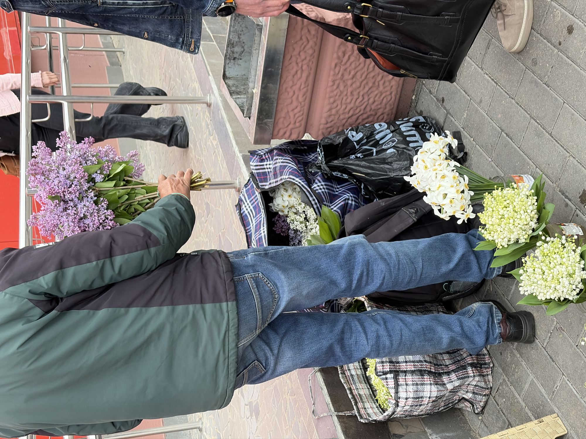 In Бараўлянскі сельскі Савет, Belarus, the image shows a street scene with various individuals and items. A man in a green and black jacket and jeans holds a bouquet of purple lilacs. Several bags, including a black plastic bag with white writing, a plaid bag, and a blue and white checkered bag, are placed on the ground. These bags contain flowers, including white daffodils and lily of the valley. Above these items, a person in a denim jacket and another holding a black handbag are visible. The background includes a metal railing, steps, and a tiled sidewalk. The scene appears to be an outdoor market or a flower vendor's setup.