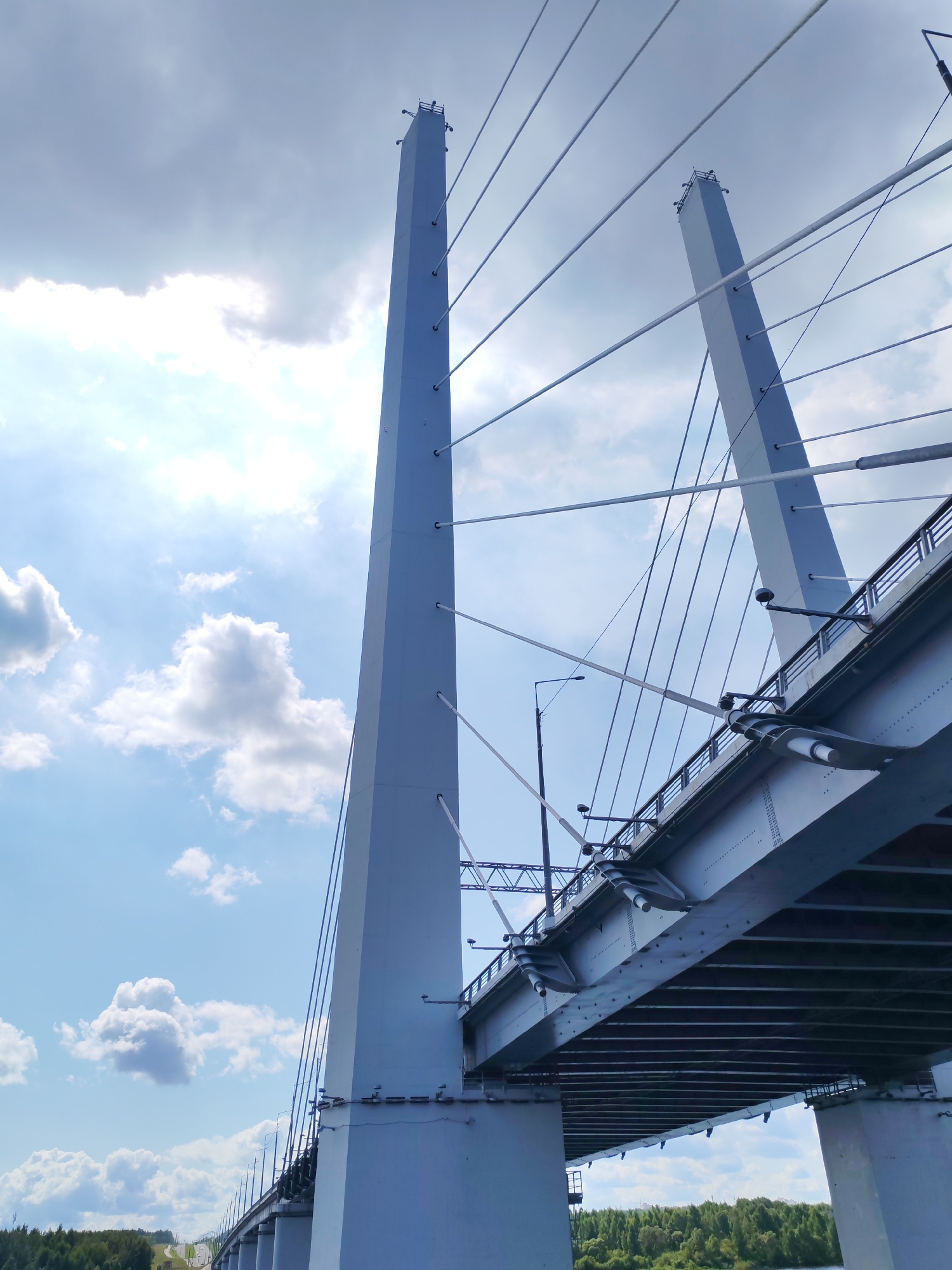 A low-angle, wide shot shows a large, modern cable-stayed bridge against a bright, cloudy sky. Two prominent, tall, light-gray towers dominate the frame, one on the left and one to the right and further back. Numerous taut, white cables extend from the tops of these towers, fanning out to support the bridge deck. The bridge itself is a dark gray structure, seen from underneath and from the side, revealing its intricate engineering with visible beams, rivets, and other structural components. Streetlights with black housings are mounted along the side of the bridge. In the background, the sky is a dynamic mix of bright blue and white clouds, with the sun peeking through, creating a bright glow. Below the bridge, a band of dark green trees forms a natural landscape, with a hint of a road and distant structures visible on the left. The water of a river is visible at the very bottom of the frame. The scene is devoid of any people or vehicles, presenting a purely architectural and environmental view.