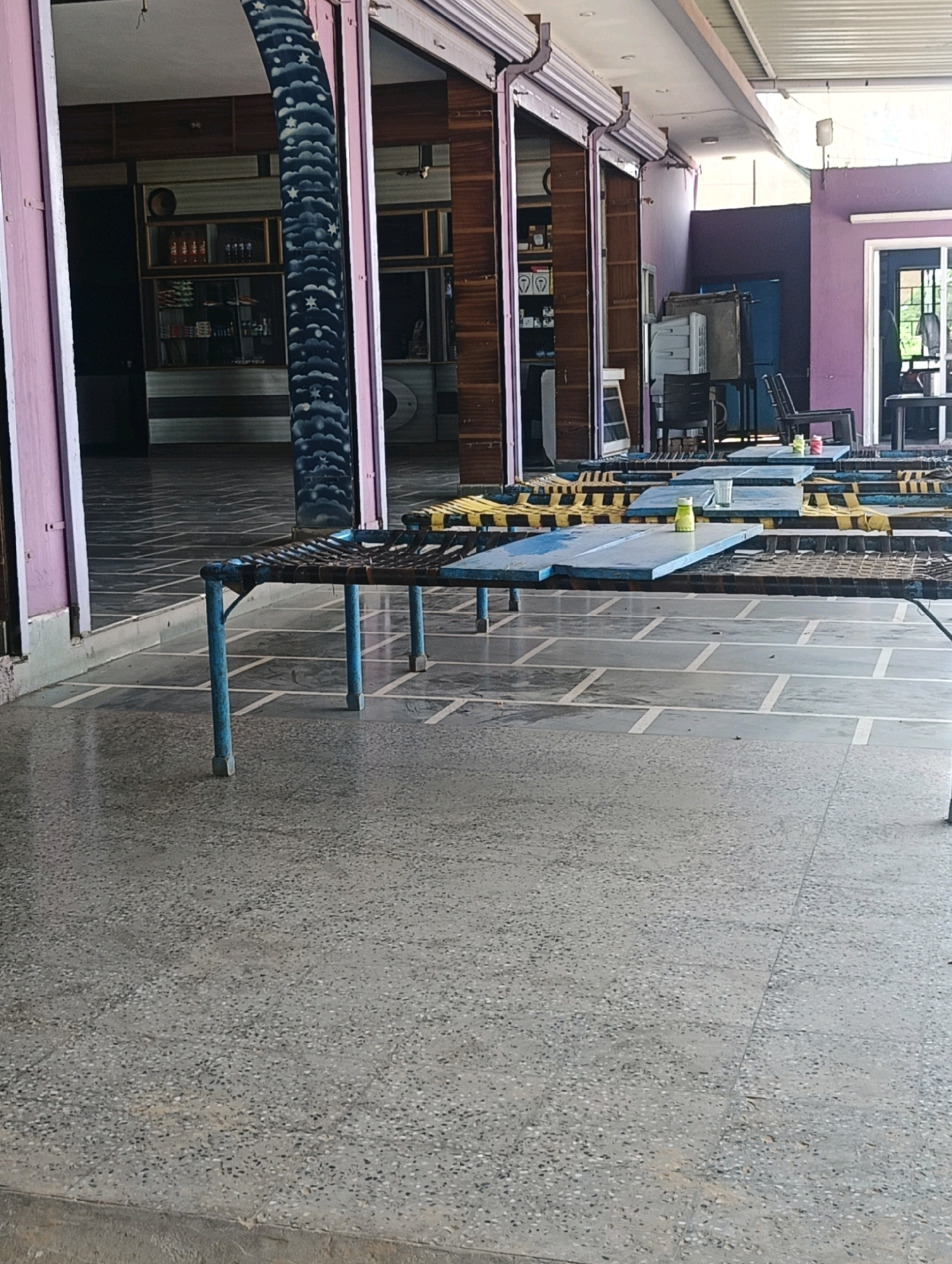 The image depicts an outdoor seating area with multiple tables and chairs, likely a restaurant or cafe, in Chhapri Bari, India. The seating arrangements are set on a paved floor with a patterned grid of light grey tiles. Several low tables, made of blue planks with visible weave-like mats underneath, are arranged in a row, supported by blue metal legs. Some tables have a yellow covering. On one of the tables, a yellow bottle and a glass are visible. In the background, a building with large open doorways and roller shutters is present. The architecture suggests a local establishment. The overall scene is empty of people.