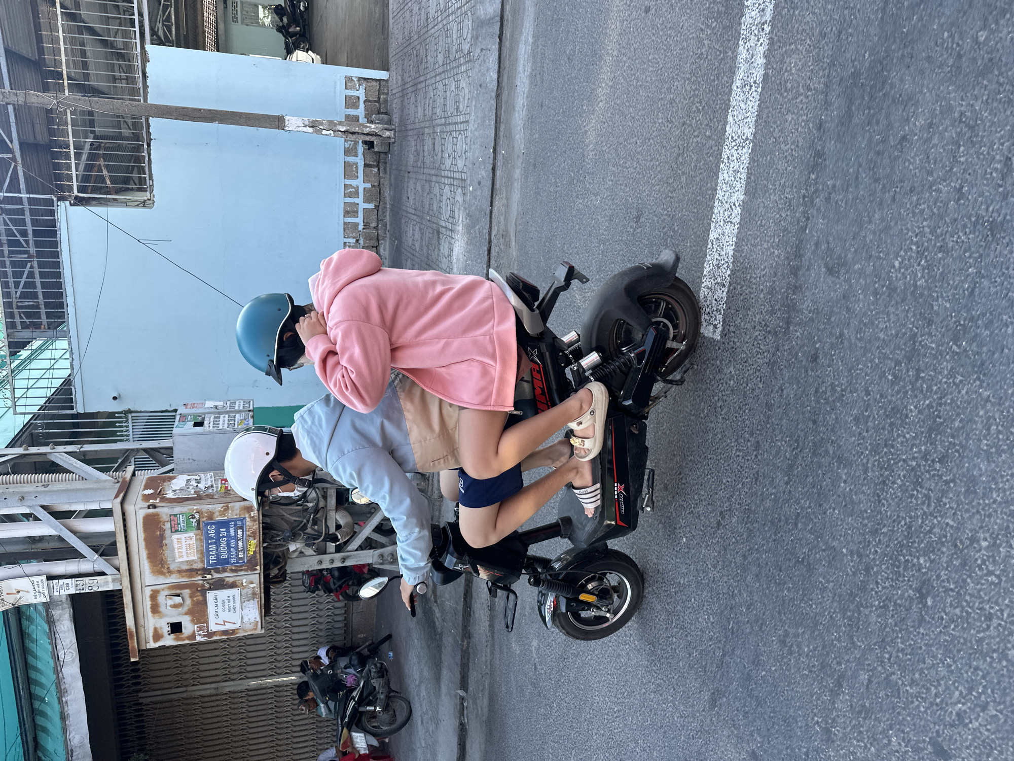 In Nha Trang, Vietnam, two individuals are riding on a black electric scooter along a paved road. The front rider, wearing a light blue jacket and a white helmet, is operating the scooter. The passenger behind, dressed in a pink hoodie and a teal helmet, is seated behind the driver, with their legs visible. The scooter displays the brand name "IMMEX" on its side. The road features a white line marking. In the background, a light blue building stands alongside other structures, including what appears to be an electrical utility box mounted on a pole with various warning signs. Several parked motorcycles are also visible.
