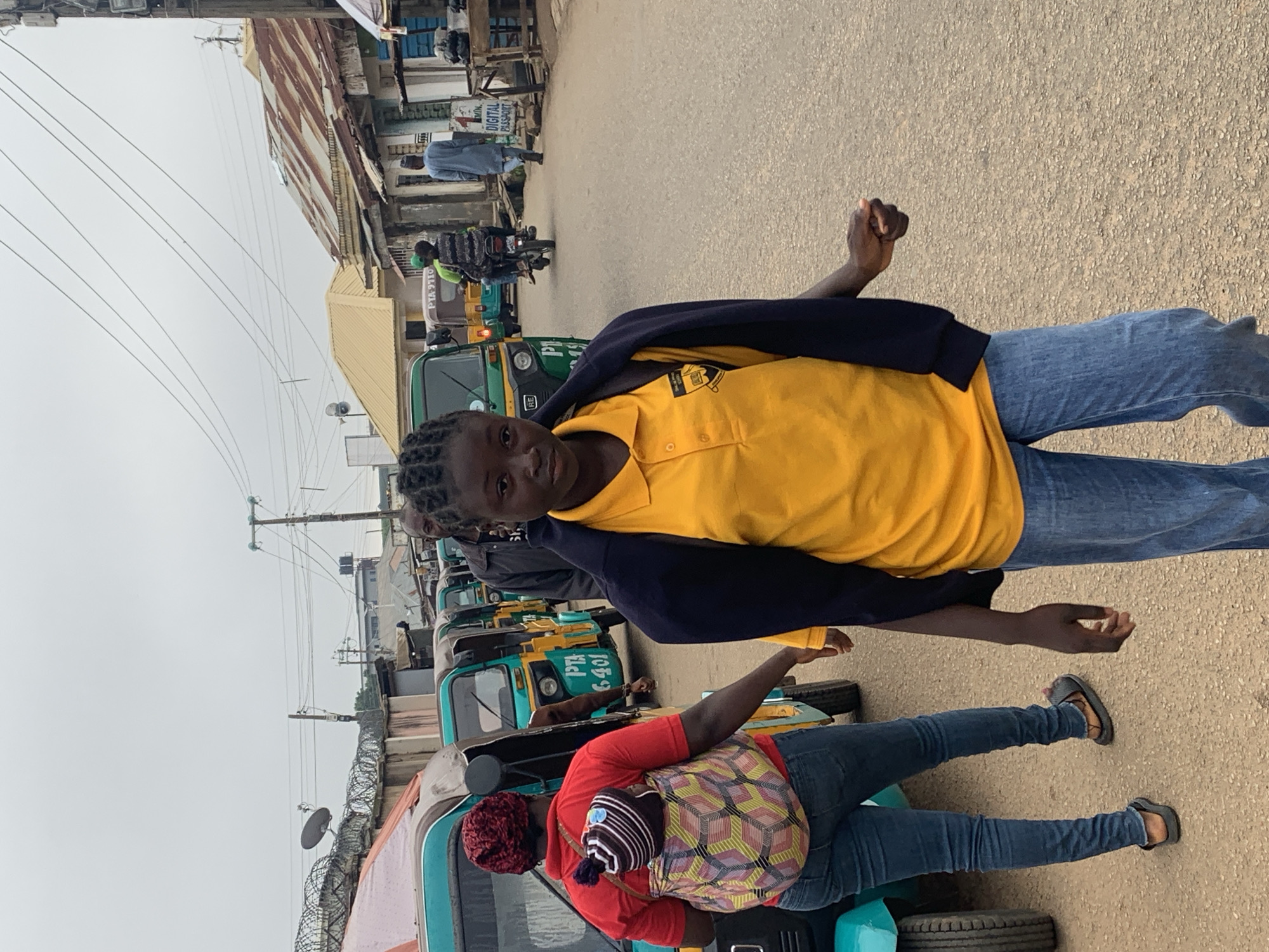 A low-angle, outdoor, medium shot captures a street scene in Jos, Nigeria, under an overcast sky. The foreground is dominated by two people. On the left, a young person with braided hair wears a yellow polo shirt and a dark blue jacket slung over their shoulders, paired with blue jeans. They stand with their left hand raised, a gesture that could imply acknowledgement or surprise. To their right, a person in a red hoodie and blue jeans is seen from the back, wearing a patterned wrap carrying a baby on their back. They are standing next to a teal-colored tricycle.
Behind them, a line of teal and yellow tricycles, commonly known as Keke Napeps, are parked on the side of a paved road. These vehicles are a primary mode of transportation in the area. The background reveals a bustling street with buildings, some featuring corrugated metal roofs and weathered facades. Electrical wires crisscross overhead, typical of many urban Nigerian settings. A few other individuals are visible in the distance, including someone riding a motorcycle and another walking. One of the tricycles has text visible: "PTB 9110" and another partially visible "P 6-401". The overall atmosphere suggests an active, everyday scene in a Nigerian town. The lighting indicates it is likely daytime, but the cloudy sky diffuses the light.