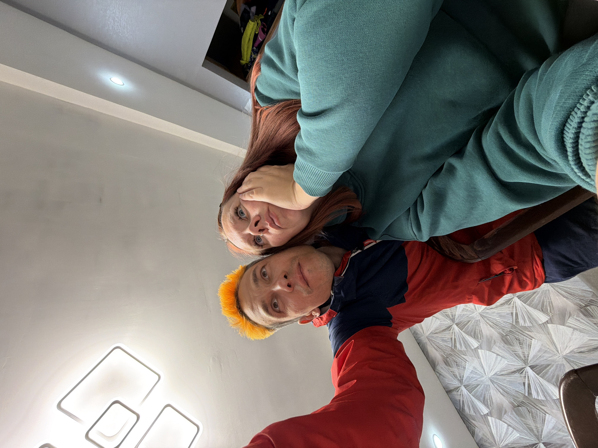 A low-angle indoor photograph captures two individuals, an adult woman and an adult man, looking directly at the camera. The image is taken from below, giving a "worms-eye view" perspective.
The woman is positioned behind and slightly above the man, her head tilted slightly to her left. She has long, reddish-auburn hair and is wearing a teal or dark green hooded sweatshirt. Her left hand is raised to her cheek, and she has light blue eyes with a somewhat serious or contemplative expression.
The man is closer to the camera, in the foreground, with short, spiky hair dyed a bright orange-yellow. He is wearing a long-sleeved top that is red on the bottom and navy blue on the top. His expression is also neutral, looking straight ahead with light-colored eyes.
The background reveals a modern indoor setting, likely a home or apartment in Chaikovsky, Russia. The ceiling is white with recessed lighting and prominent, contemporary square-shaped light fixtures that emit a bright white glow. Part of a dark shelving unit or closet, containing colorful clothing, is visible behind the woman. To the right, a wall features a distinctive grey and white geometric patterned wallpaper, resembling abstract fans or starbursts. A dark brown piece of furniture, possibly the back of a chair, is partially visible on the far right. The lighting appears to be entirely artificial, suggesting either evening or an interior space relying solely on electric lights. No text is visible in the image.