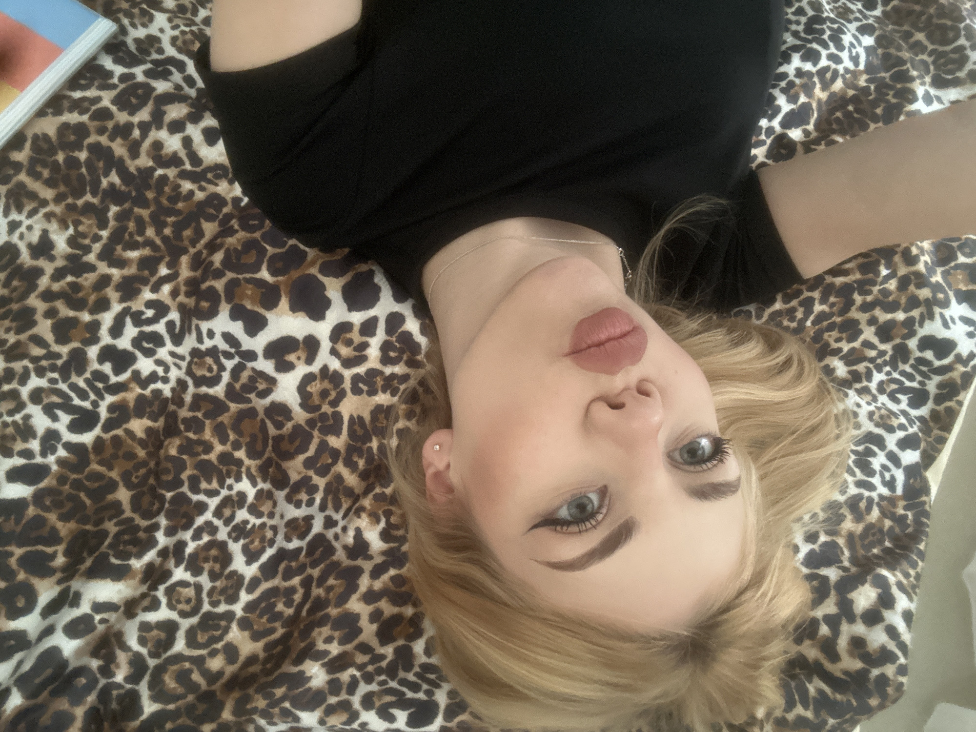 A young woman with blonde hair and blue eyes is lying on a bed with a leopard print sheet. She is wearing a black shirt. The location is Unknown, Unknown.