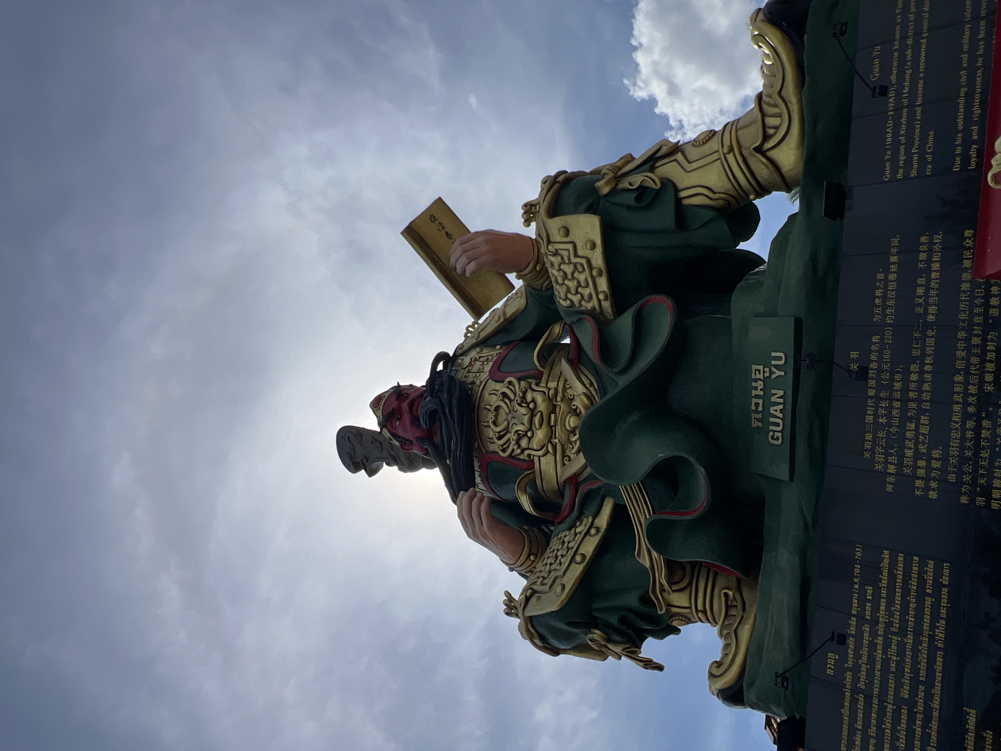 An outdoor, eye-level, full-shot, low-angle photograph depicts a large, seated statue of Guan Yu against a bright, cloudy sky. The statue is elaborately decorated with gold accents against a dark green robe and armor. Guan Yu's face is painted red, with a long black beard and stern expression. He holds a golden book in his right hand and a weapon behind his head. The base of the statue has an inscription in Thai and English. The English inscription reads: "Guan Yu (160 AD-219 AD), otherwise known as Yun Chang, was a general of the state of Shu Han in the Three Kingdoms period of China. He was born in Xieyu County, Hedong (a sub-district of present-day Yuncheng in Shanxi Province) and became a renowned general during the Three Kingdoms era of China. Due to his outstanding civil and military talents, his loyalty and righteousness, he has been revered as the God of War. The statue is located in Baan Hua Thanon, Thailand." The lighting suggests it is daytime, possibly late morning or early afternoon, with the sun partially obscured by clouds and positioned behind the statue.