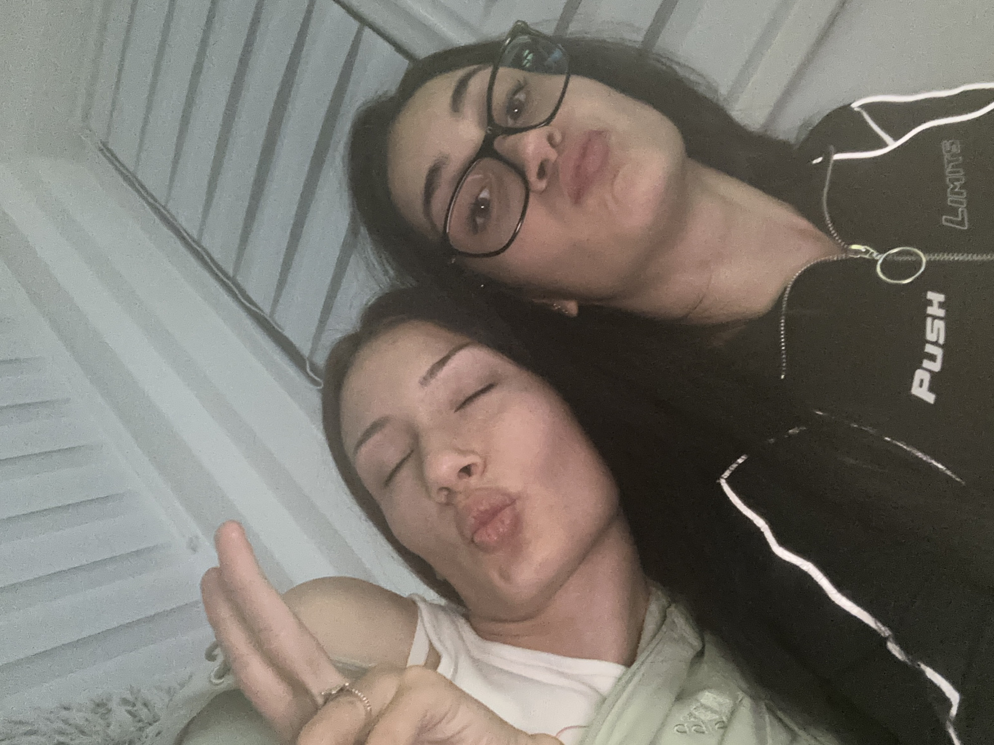 Two women are posing for a selfie indoors. The woman in the foreground has her eyes closed and is making a kissing face while holding up two fingers. She is wearing a light-colored shirt and a ring on her finger. The woman in the background is wearing glasses and also making a kissing face. She is wearing a black jacket with white accents and the words "PUSH LIMITS" visible on the jacket. The background appears to be a wall with vertical lines. The location in Burgas, Bulgaria, cannot be determined from the visual information provided.