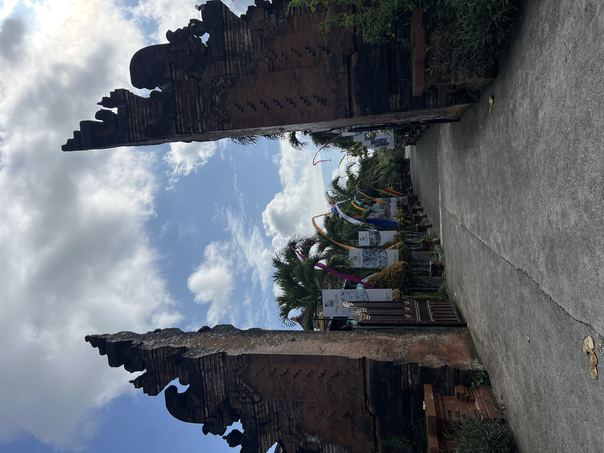 The image captures a pathway flanked by traditional Balinese brick structures in Ubud District, Indonesia. The sky is visible between the structures, showing a mix of blue sky and white clouds. Decorations, including colorful ribbons and banners, adorn the pathway. The ground is a paved surface. The photo appears to have been taken during the daytime, with natural lighting.