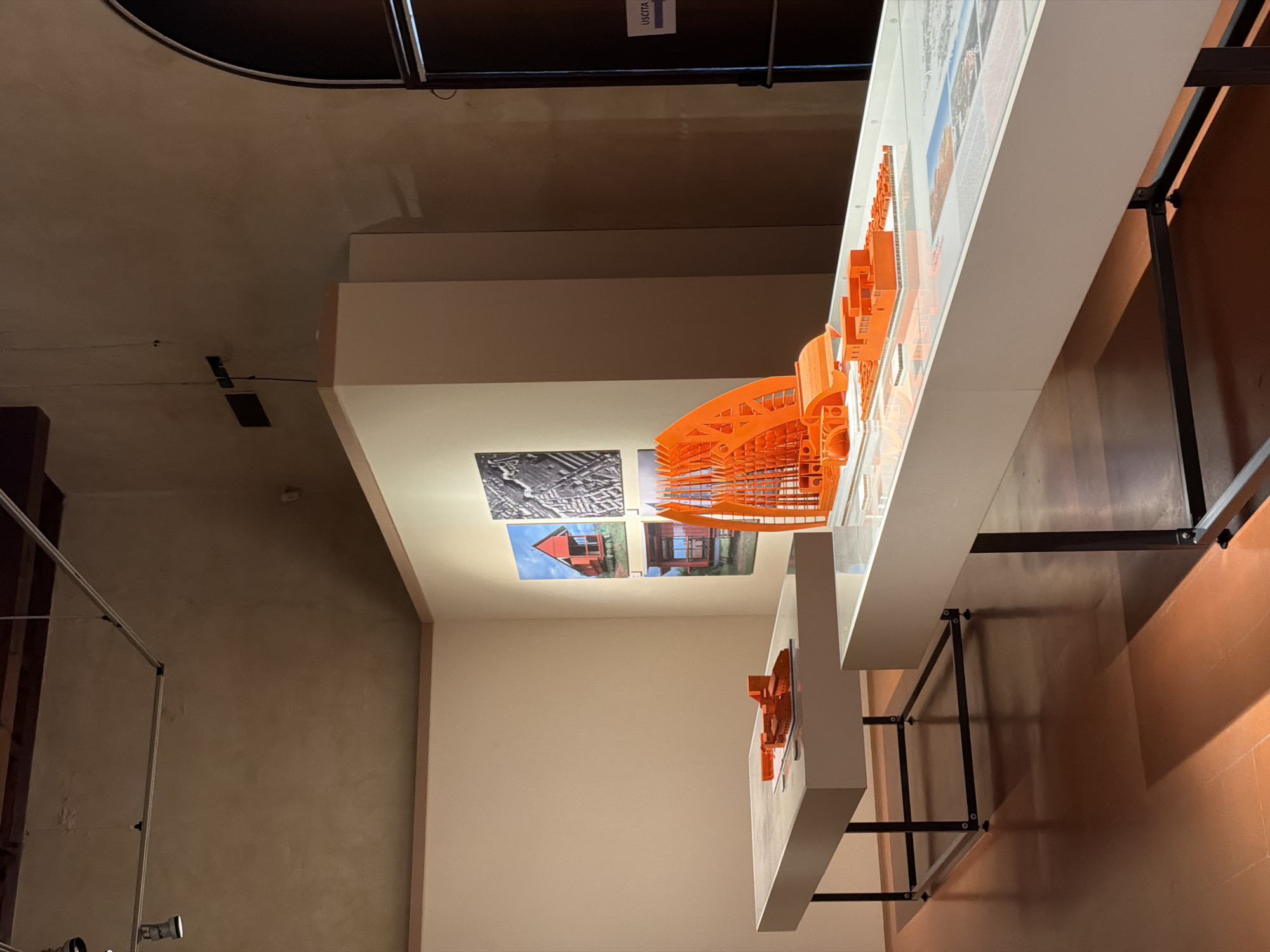 This image shows an indoor exhibition of architectural models in Mantua, Italy. There is an orange architectural model of a building on a white table, with architectural plans and pictures behind it, all mounted on a white wall. A second model is on a lower white table, further in the background. The room has neutral-toned walls and a dark ceiling with some suspended lighting structures. The floor is a glossy brown surface. The room appears to be a gallery or museum space.