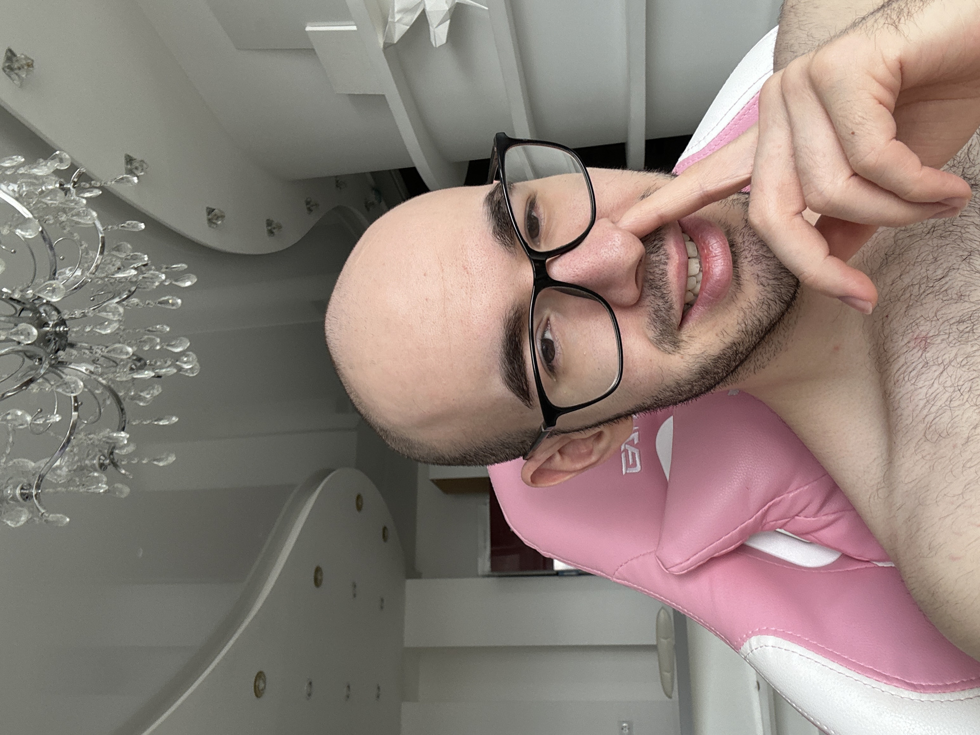 The image, rotated 90 degrees clockwise, presents a close-up of a man posing in an indoor setting. The subject is a bald man with dark stubble or a short beard, wearing black-rimmed glasses. He has dark eyes and is seen smiling or smirking, revealing his teeth and pink lips. He appears bare-chested, with body hair visible on his chest and arm. His right hand is raised, with his index finger touching his nose in a seemingly playful gesture, while his other fingers are extended.
The man is seated in a pink and white gaming chair; the backrest, which features the letters "EA" (or similar, possibly "EVA" with the 'V' partially obscured) in white, and part of the seat are visible. The background consists of a modern interior with white walls and a white ceiling that features curved, layered architectural details and small, square, crystal-like decorative elements. An ornate crystal chandelier with numerous clear drops and a silver frame hangs prominently on the left side of the image. Further in the background, a white piece of furniture with circular cutouts is partially visible. The lighting is bright, consistent with an indoor setting, and the overall atmosphere is casual and cheerful. The scene is set in Khimki, Russia.
