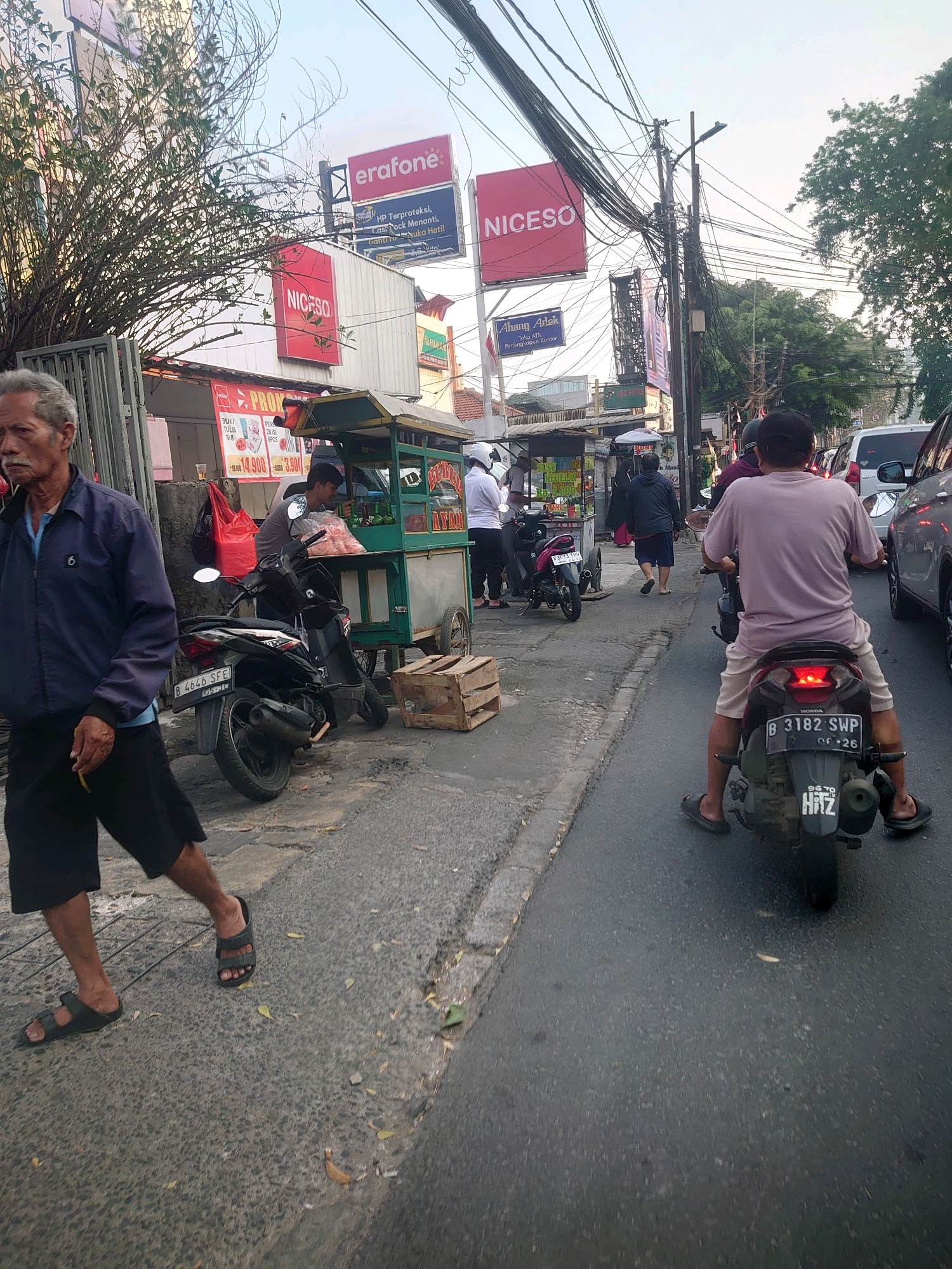 The image depicts a street scene in South Jakarta, Indonesia, characterized by heavy traffic and commercial activity. On the left side of the frame, an older man in a blue jacket and black shorts walks along a sidewalk, holding a cigarette. Adjacent to him is a parked black motorcycle with license plate B 4646 SFE. Behind the motorcycle, a food stall with a green awning is visible, selling what appears to be fried chicken (Ayam Goreng). A person in a gray shirt is attending the stall. Further down the street, several more motorcycles are parked, and people are milling about.
The background is dominated by a cluttered overhead network of electrical wires and poles. Several prominent billboards advertise various businesses, including "Erafone" and "NICESO." The road itself is asphalted and shows signs of wear. A motorbike with a rider wearing a pink shirt and light-colored shorts is in the foreground, moving away from the viewer. The motorcycle's license plate is B 3182 SWP. Traffic is moderate, with other vehicles, including cars, visible in the distance. The sky is overcast, suggesting it might be late afternoon or early evening. The overall impression is of a busy urban environment with a mix of residential and commercial elements.