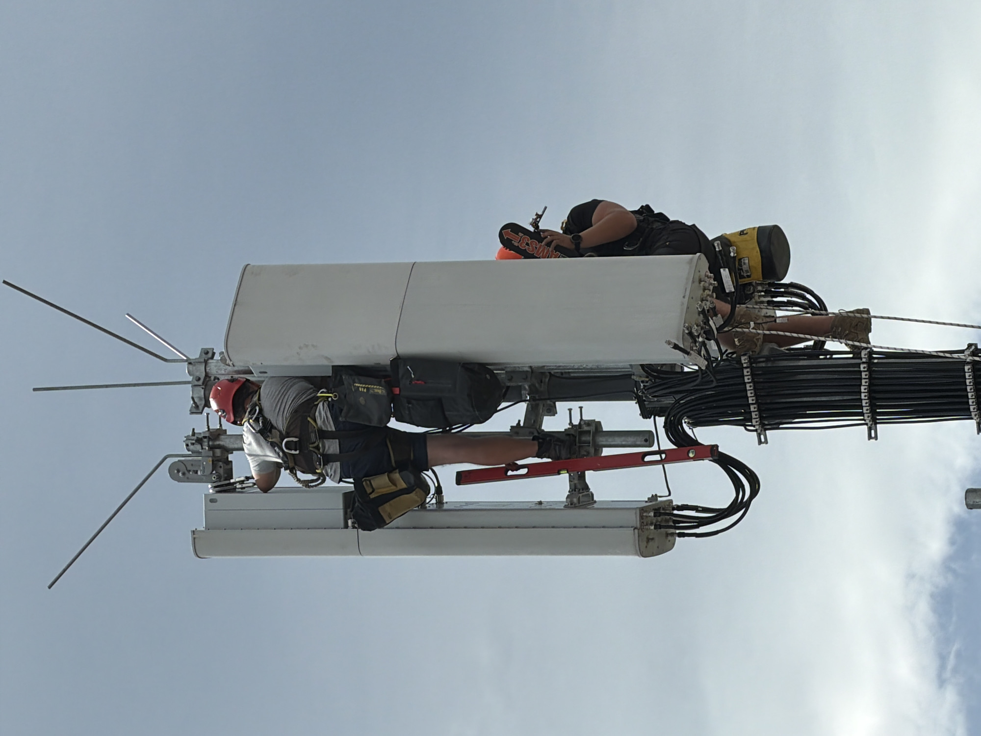 Two workers in safety harnesses and hard hats are performing maintenance on a cell tower against a pale blue sky with scattered clouds. The worker on the left is higher up, using a red level as they work on the tower's equipment. They are wearing a grey shirt, dark shorts, and a red helmet. The worker on the right is below and to the right of the first worker, holding a tool and wearing a black t-shirt and orange helmet. Both workers have tool pouches attached to their harnesses. The cell tower has large white rectangular panels and is covered in black cables. The image does not provide any specific information about the location within the city of Munich, Germany.