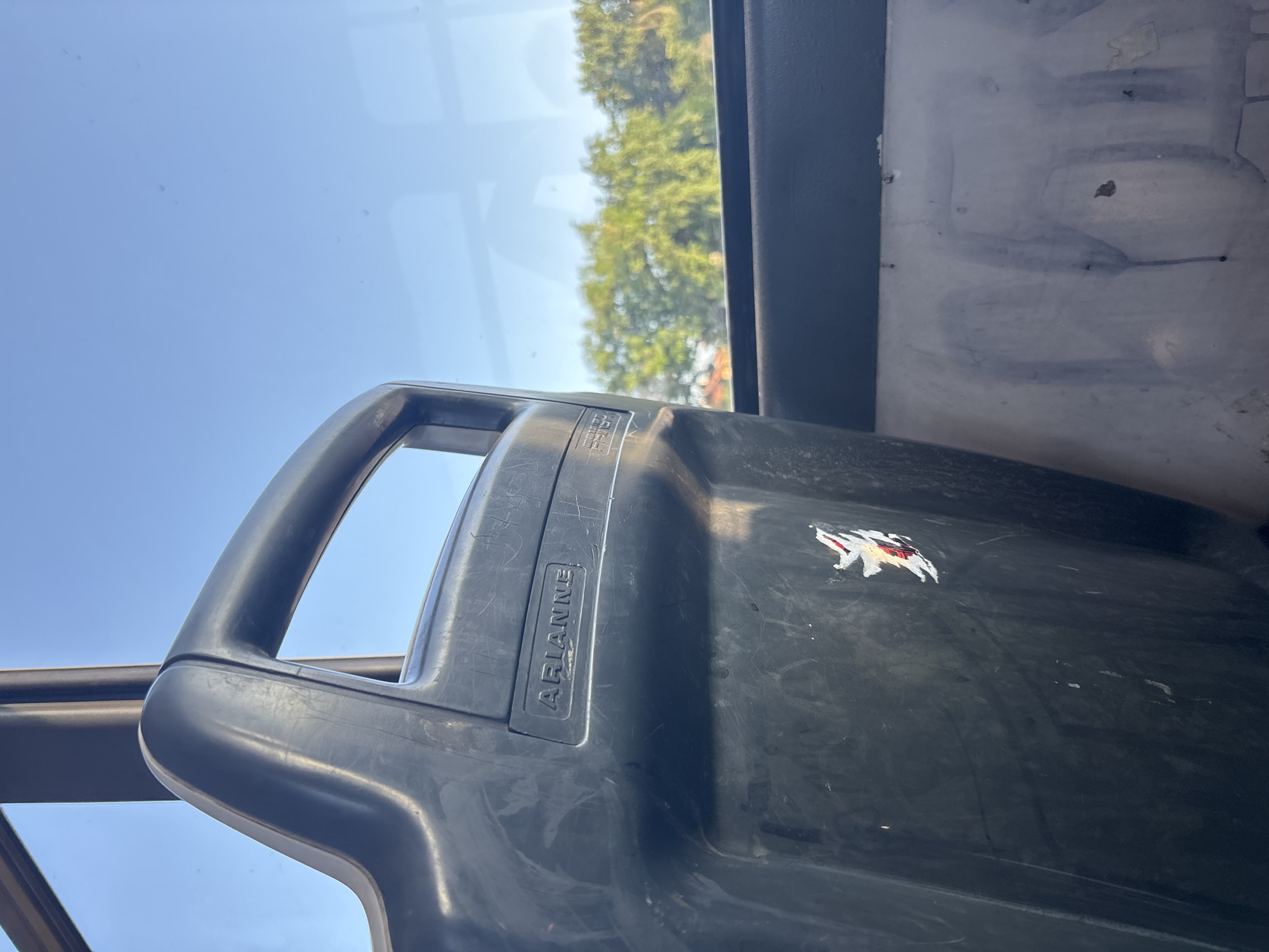 The image captures an angled view from inside a vehicle, looking towards a window. A dark, possibly plastic, seat or container with a prominent handle occupies the foreground and center, extending diagonally across the frame. The surface of this object shows numerous scratches and wear. Embossed on its side, the word "ARIANNE" is clearly legible. A distinctive white and red sticker, depicting a stylized animal (possibly a wolf or bear) with red accents, is affixed to the right side of the dark object.
Through a clear window in the upper part of the image, a bright, cloudless blue sky is visible, indicating a sunny day. Below the sky, out the window, blurred green foliage from trees can be seen, suggesting an outdoor, natural setting. To the right of the dark object, an interior wall or panel is visible, appearing worn with smudges and faint outlines. No people are present in the scene, and no specific activity is occurring.