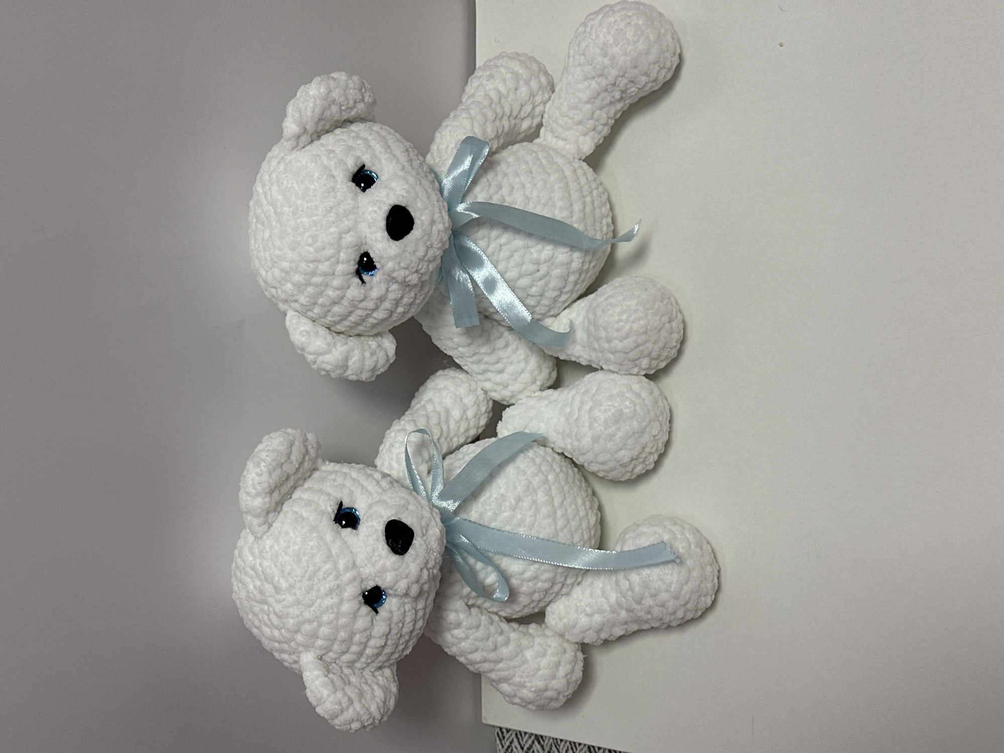 Two crocheted white teddy bears sit side-by-side on a white surface against a plain grey background. Both bears have blue eyes, black noses, and light blue satin ribbons tied in bows around their necks. The bear on the left is positioned slightly more towards the viewer, with its legs extended forward. The bear on the right is angled slightly away, with its legs tucked closer to its body. The texture of the crochet is visible on the bears' bodies, suggesting a handmade quality. The lighting is even, with a slight shadow cast behind the bears. There is no indication of the city of Ufa, Russia, or any specific location within it.