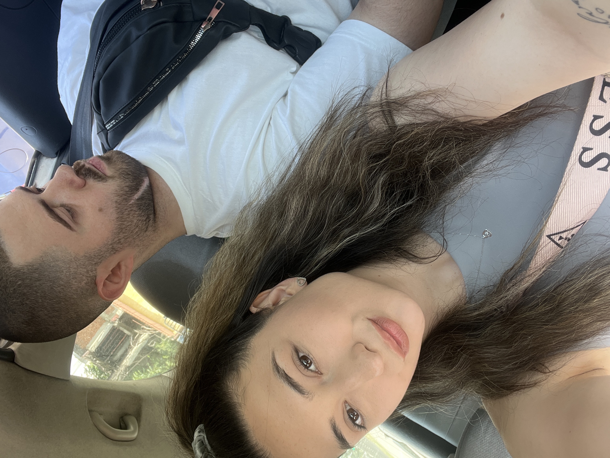 A medium shot captures a young couple in a car, likely in Plovdiv, Bulgaria, based on the architectural reflections in the window. The woman, in the foreground, faces the camera, her long brown hair cascading over her shoulder. She wears a grey top and a delicate necklace. To her right and slightly behind, the man is seen in profile, his head tilted to the side. He has short hair and a beard and is wearing a white t-shirt and a black fanny pack across his chest. The car's interior is visible, with a seatbelt across the woman and the man's arm resting on his lap. The background through the car window suggests an urban setting with buildings and trees.