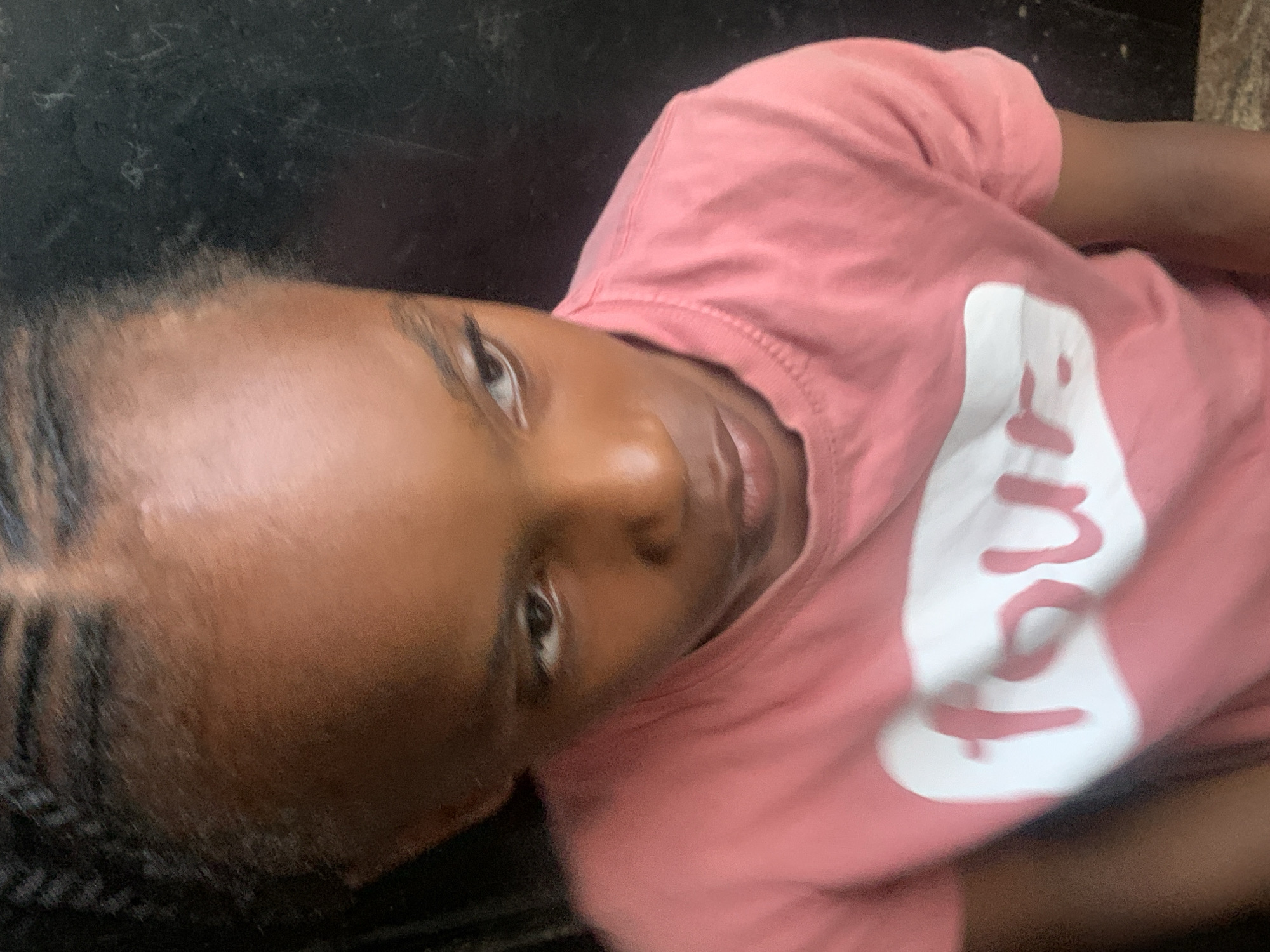 The primary image features a young person, likely a child or young adolescent, with dark skin and dark eyes, looking directly at the camera with a neutral expression. The image is oriented on its side, showing the individual's head tilted towards the viewer. Their hair is styled in dark, intricate braids, pulled back from the forehead.
The subject is wearing a short-sleeved, pink t-shirt. A significant detail is the white, stylized text printed vertically on the front of the shirt, which appears to spell "Love." The letters have a distinctive, rounded, and slightly blob-like design.
The background is uniformly dark and indistinct, offering no specific details about the surroundings or time of day. No particular activity is visible other than the subject posing for the photograph. There are no explicit location cues within the image itself, but the context provided places the scene in Barakin Akawo, Nigeria.