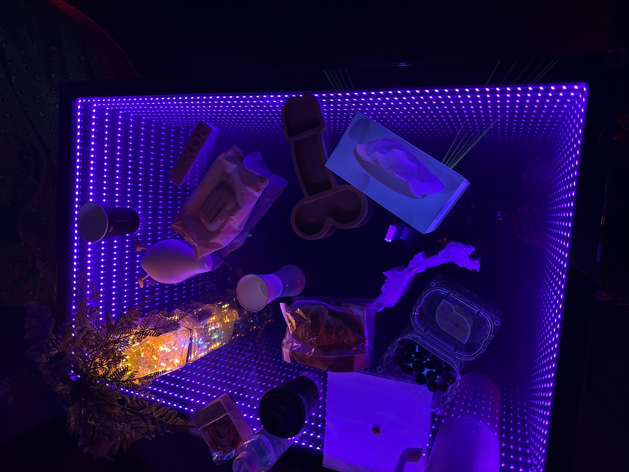 The image shows a rectangular table with a black frame. The table's surface is illuminated with a grid of closely spaced purple LED lights, creating an infinity mirror effect. A variety of items are scattered across the table including paper cups, packaged food, a wooden object shaped like male genitals, a tissue box, a plastic container with fruit, a gift with foliage, and a water bottle. The setting appears to be indoors, in a dark environment with the light from the table being the primary illumination. The location and city cannot be determined from the image.