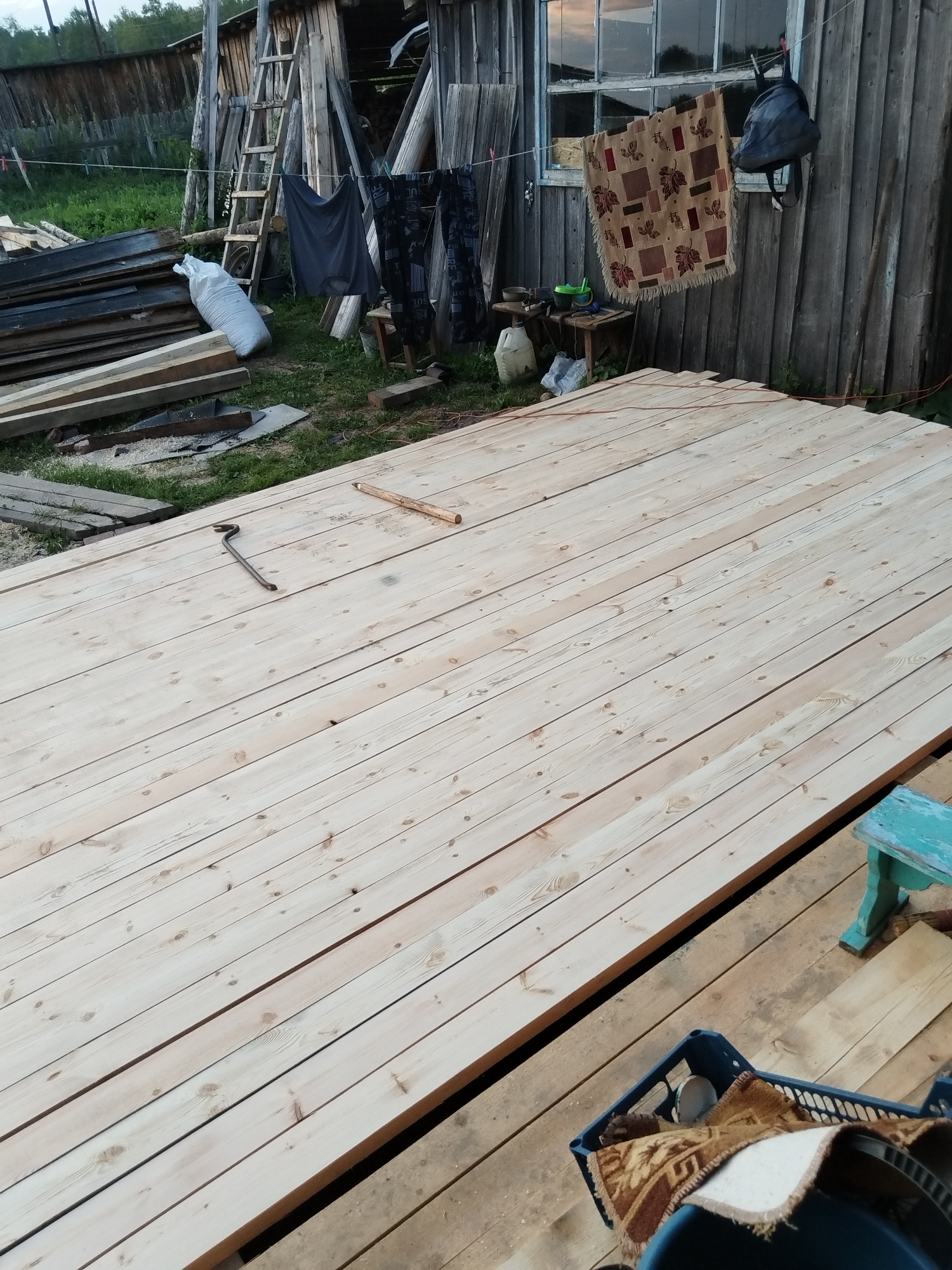 A sunlit outdoor scene in Minayevka, Russia, shows a partially constructed wooden floor made of light-colored planks. A crowbar lies on the planks, along with a short piece of wood. In the background, a weathered wooden structure with a ladder leaning against it stands amidst scattered lumber and gardening tools. A rug and clothing hang from a clothesline in front of the structure. The ground is covered in grass and dirt.