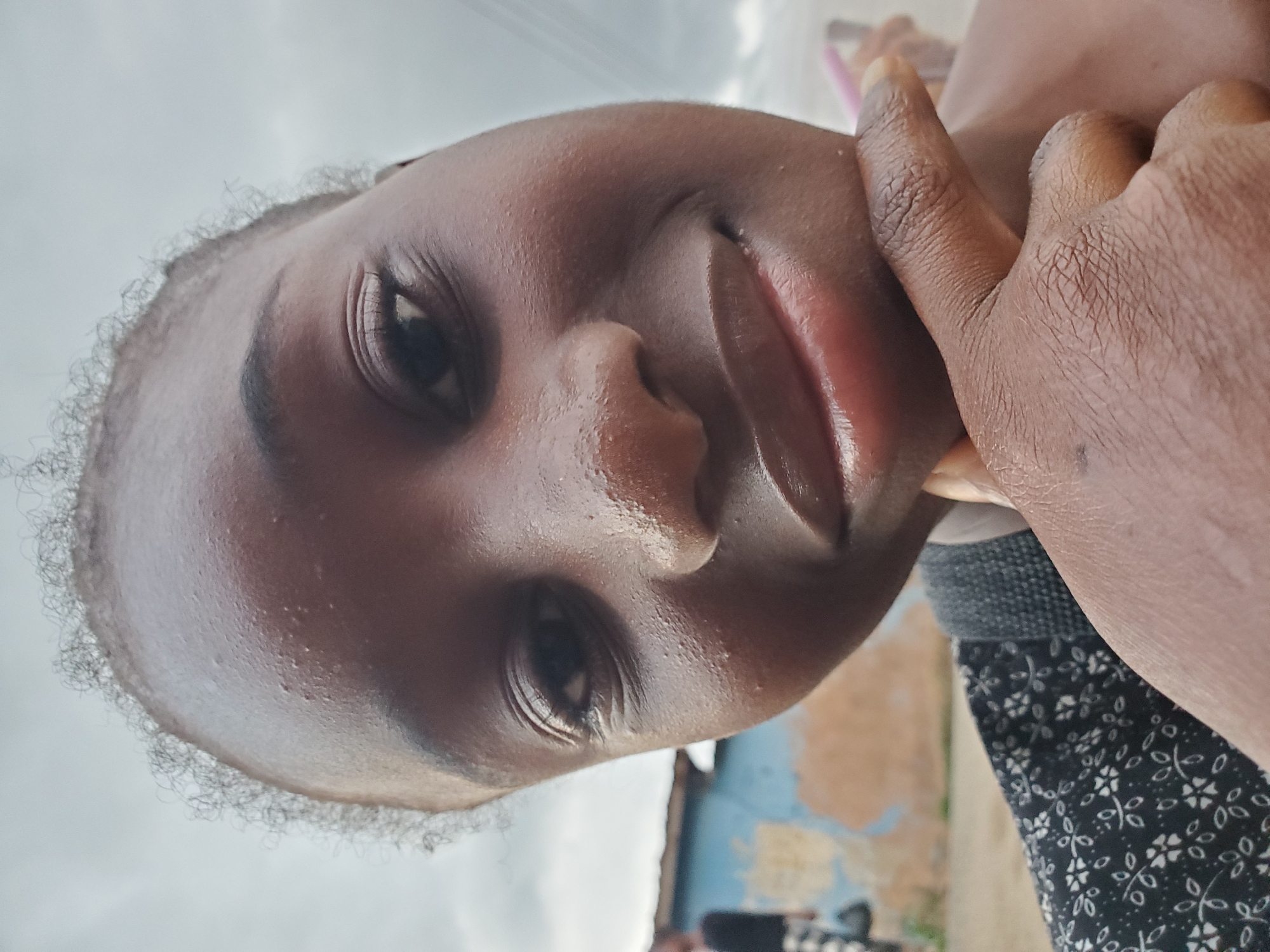 This is a close-up, upward-facing portrait of a young Black person. Their face is dominant in the frame, with the top of their head and a hand reaching towards their face also visible. The subject has dark skin and appears to be looking upwards. The lighting suggests it could be daytime, possibly overcast, as the sky is bright but lacks harsh shadows.
The person's hand, with visible skin texture and fingernails, is gently touching their cheek and chin area, partially obscuring their jawline. Their lips are slightly parted, and their eyes are open and looking upward. Some stray curly hair is visible at the edge of their forehead.
In the background, there are hints of an outdoor setting. A blurry, textured wall with patches of blue paint is visible, along with what appears to be another person's clothing with a floral pattern. This suggests a casual, possibly residential environment. The overall mood is serene and perhaps a bit contemplative. There is no visible text in the image. The location context is Jos, Nigeria.