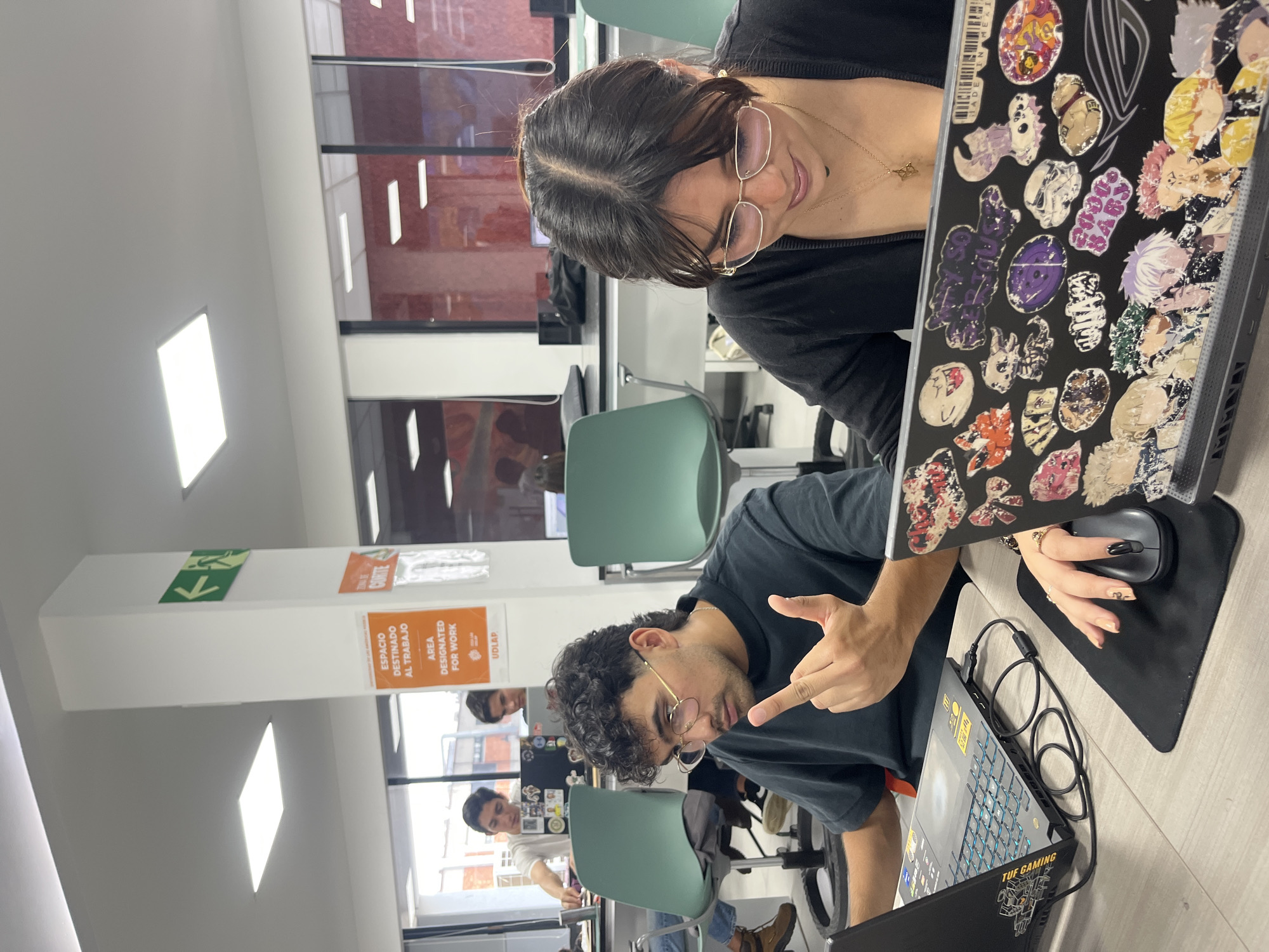 Two people are at a desk working on laptops in a brightly lit area. A woman with glasses and a black top is looking at the laptop with stickers on it; her hand is on a black mouse. A man is in front of her with a black shirt and glasses. He is pointing with his right hand. The laptop in front of him has "TUF GAMING" visible on the front. In the background, there is an area designated for work sign with the words "Espacio destinado al trabajo Area Designated for Work" along with the logo of the Universidad de las Américas Puebla (UDLAP) visible in the background, placing the location in San Andrés Cholula, Mexico. Other people are visible in the background working at desks. A green exit sign is also visible.