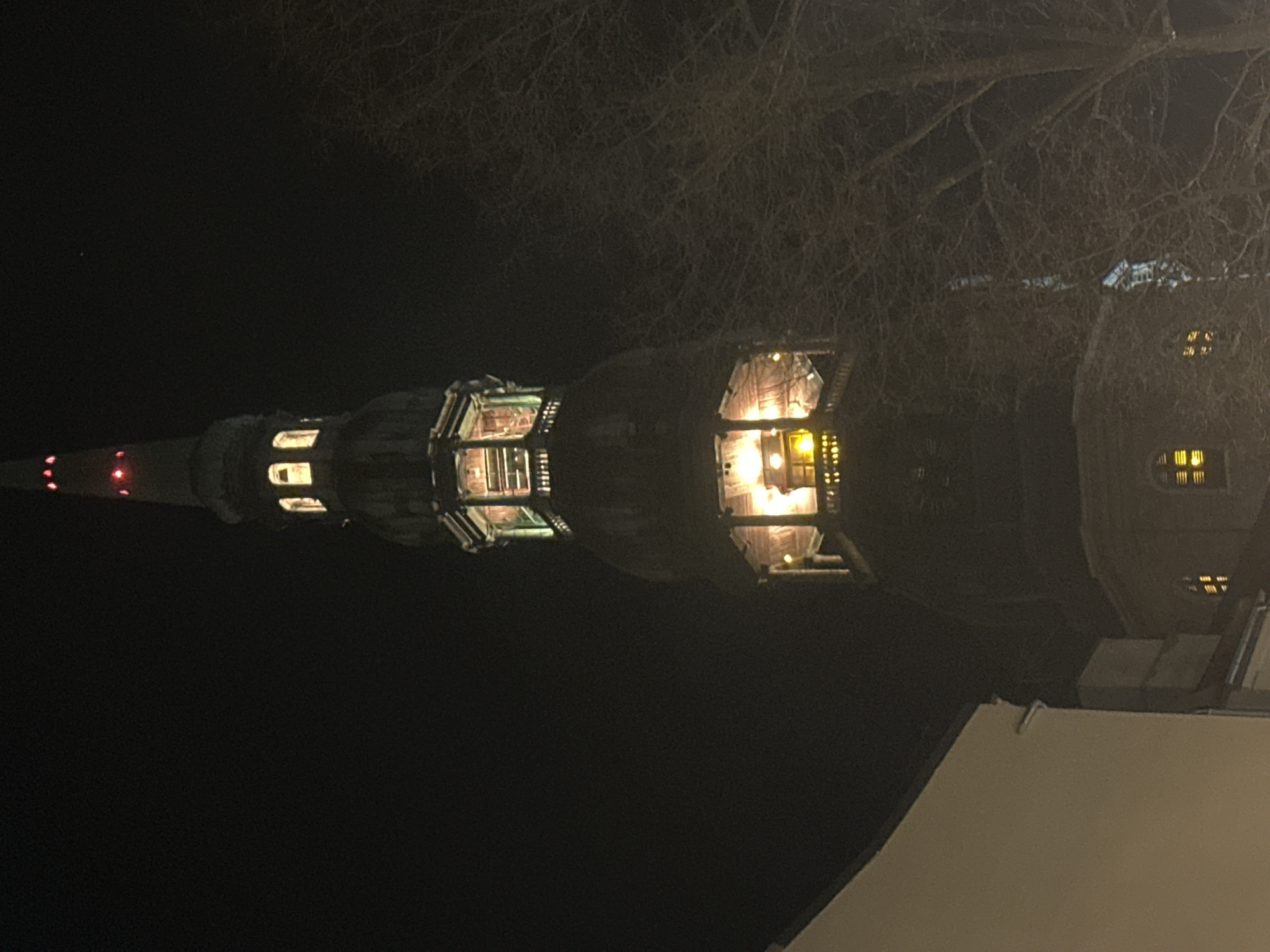 At night, the illuminated spire of St. Peter's Church in Riga, Latvia, dominates the view. The spire, with its distinctive tiered structure, is visible in the dark sky, punctuated by light emanating from its windows and culminating in a red-lit tip. To the right of the spire, a darker, rounded building is partly obscured by leafless tree branches. The lower part of the frame is occupied by a light-colored, flat rooftop.