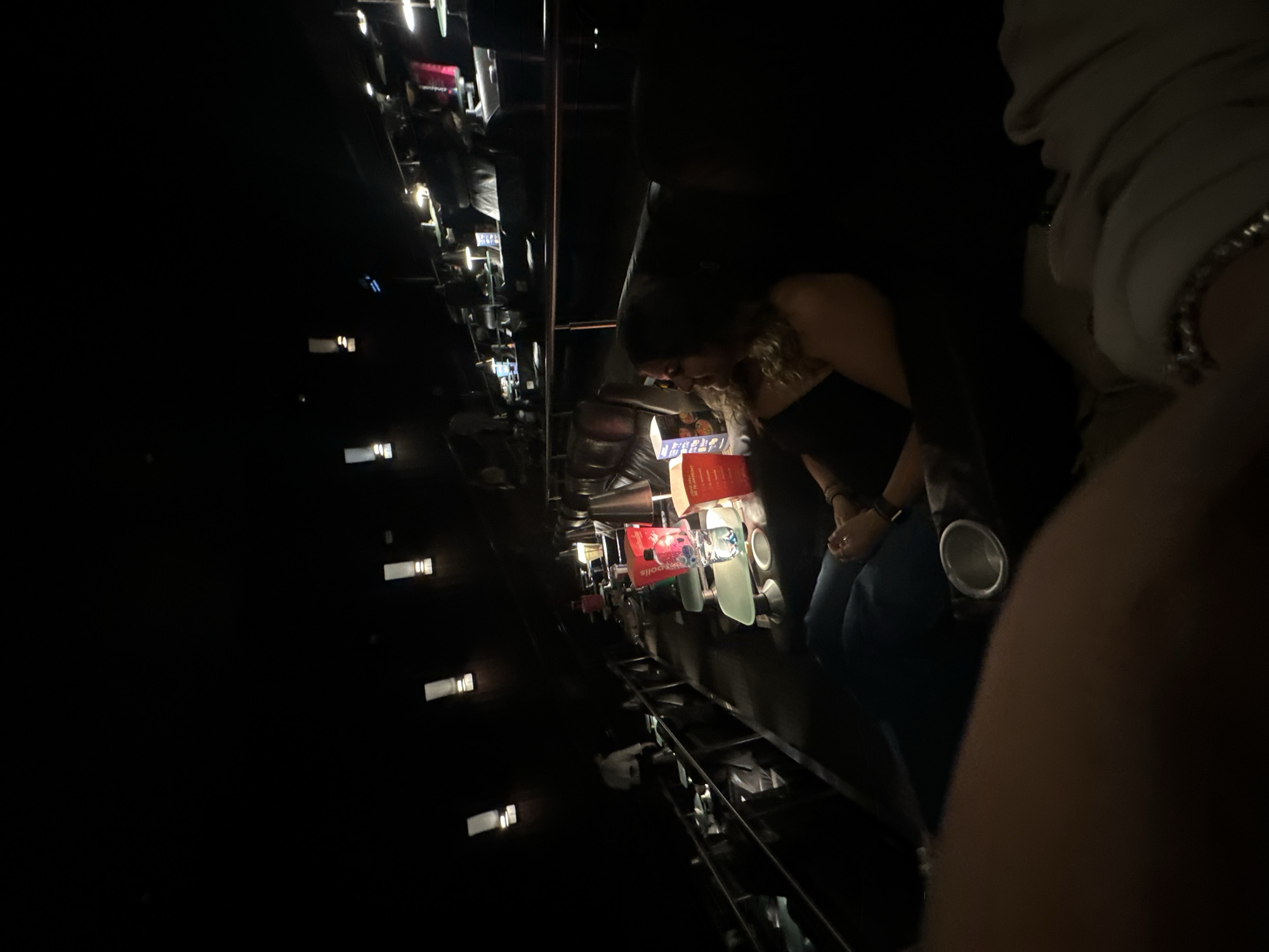 Captured in what appears to be a dark setting within Huixquilucan de Degollado, Mexico, the image features a group of individuals, with one person positioned closer to the camera appearing to examine or interact with objects below, which appear to be refreshments, possibly at a bar or event. The environment seems illuminated by artificial lighting, including ambient light from nearby sources, contributing to the overall low-light setting.