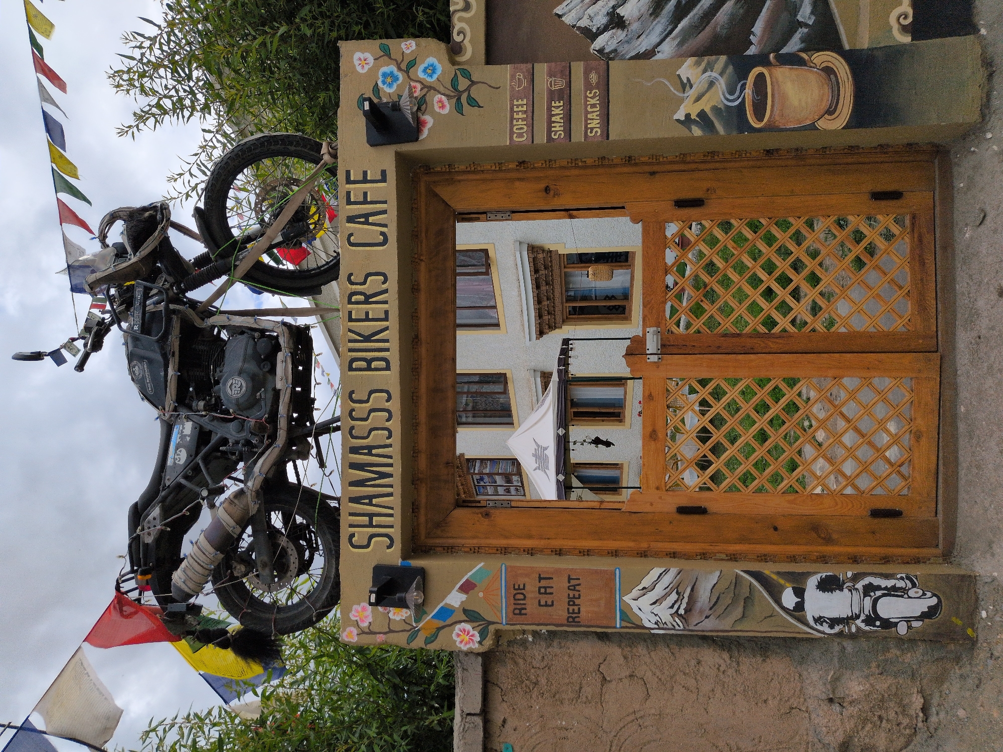 A vintage-style motorcycle is prominently displayed on top of an arched doorway entrance. The motorcycle is dark colored with chrome accents and adorned with prayer flags. The doorway is made of distressed wood with a diamond-patterned lattice on its doors, which are partially open. Above the doorway, the text "SHAMASSS BIKERS CAFE" is painted in bold, golden letters. To the left of the entrance, there are painted flowers, a small lamp, and prayer flags. Below the entrance, on the left side of the wall, is a painting of a motorcyclist riding towards a mountain landscape. On the right side of the entrance, there are painted signs for "COFFEE", "SHAKE", and "SNACKS", accompanied by small icons. Further to the right, a painting depicts a steaming cup of coffee in a mountainous setting. The building behind the doorway appears to be a guesthouse or cafe, with windows and a white tent structure visible. The overall scene suggests a biker-friendly establishment in a mountainous region, likely Leh, India, given the presence of prayer flags common in Himalayan regions. The sky is overcast.