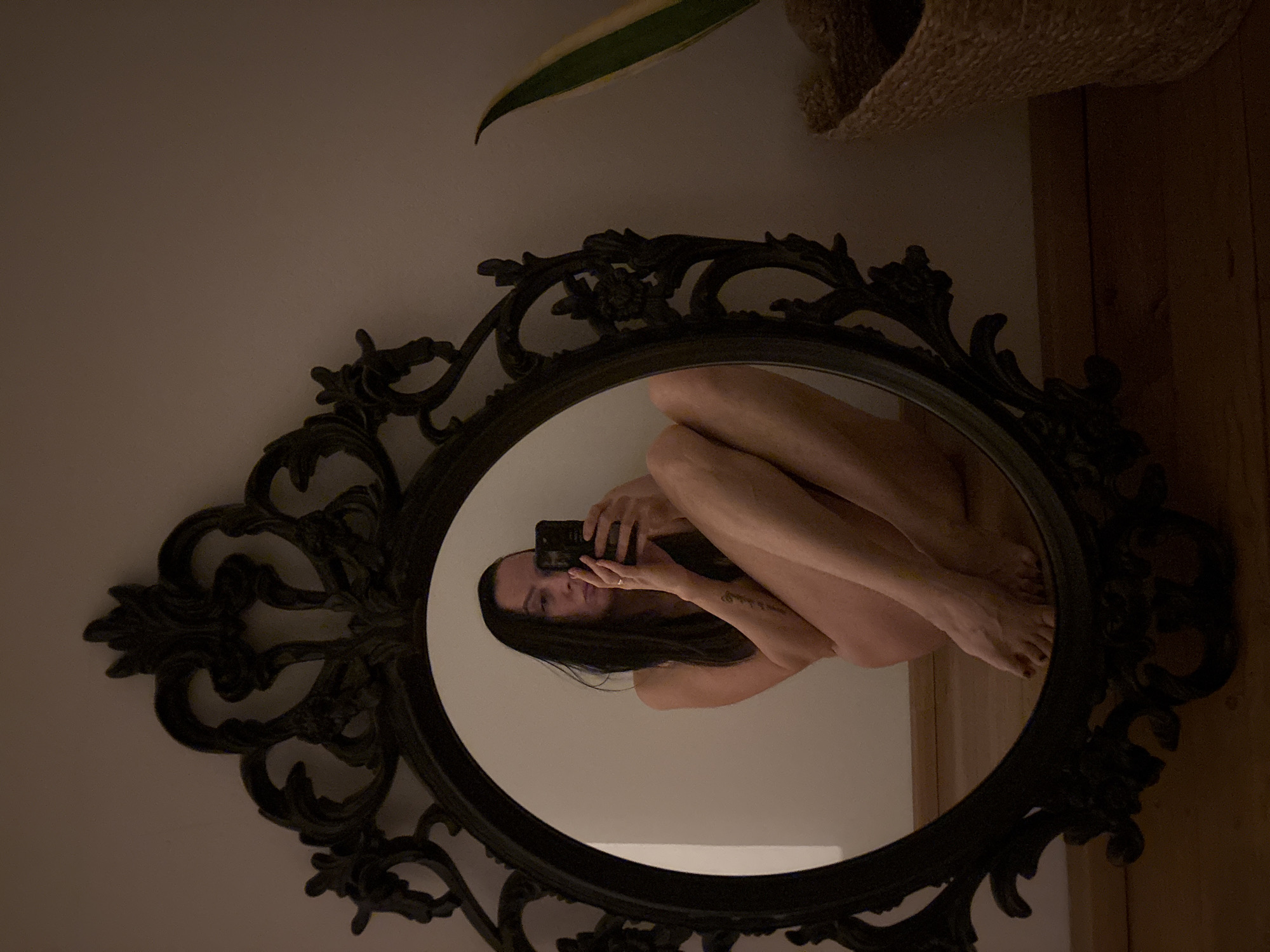 A nude woman is reflected in an oval, ornate, antique-style mirror with a dark frame. She is seated with her knees drawn up to her chest, holding a smartphone in her hands to take the picture. Her dark hair falls around her face and shoulders. The background behind the mirror includes a wall with a plant and wooden paneling. The location cannot be determined from the given information.