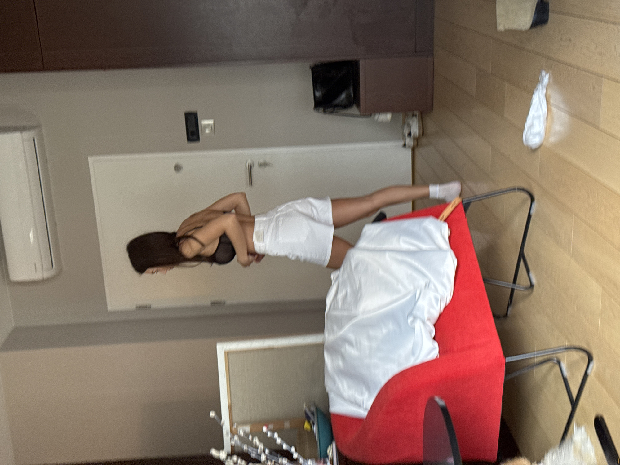 The image shows a woman wearing a black bra and white shorts, lying on a red sofa covered with a white sheet. The background includes a door, air conditioning unit, a TV and a wall unit, and a wooden floor with some objects scattered around. The specific location and city are unknown.