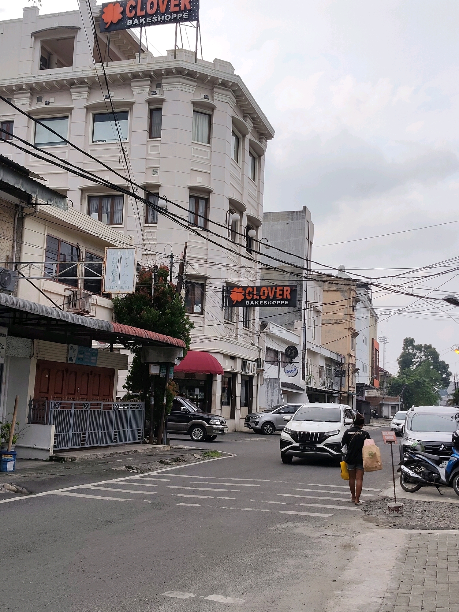 The image depicts a street scene in the City of Medan, Indonesia. In the foreground, a paved road with white crosswalk markings runs diagonally across the frame. A person with dark hair, wearing a black t-shirt with "INDONESIA" written on the back, shorts, and sandals, is walking across the crosswalk carrying two shopping bags. To the right, a parked motorcycle and a white SUV are visible. Further down the street, other vehicles, including a white SUV and a black SUV, are present.
Dominating the background is a multi-story building with ornate architectural details and a sign at the top reading "CLOVER BAKESHOPPE". Below this sign, another building with a red awning is visible, and further to the left, a building with a gray awning and wooden doors is present. Electrical wires are strung across the scene, creating a tangled network above the street. The sky is overcast. The buildings suggest a commercial or mixed-use urban environment.