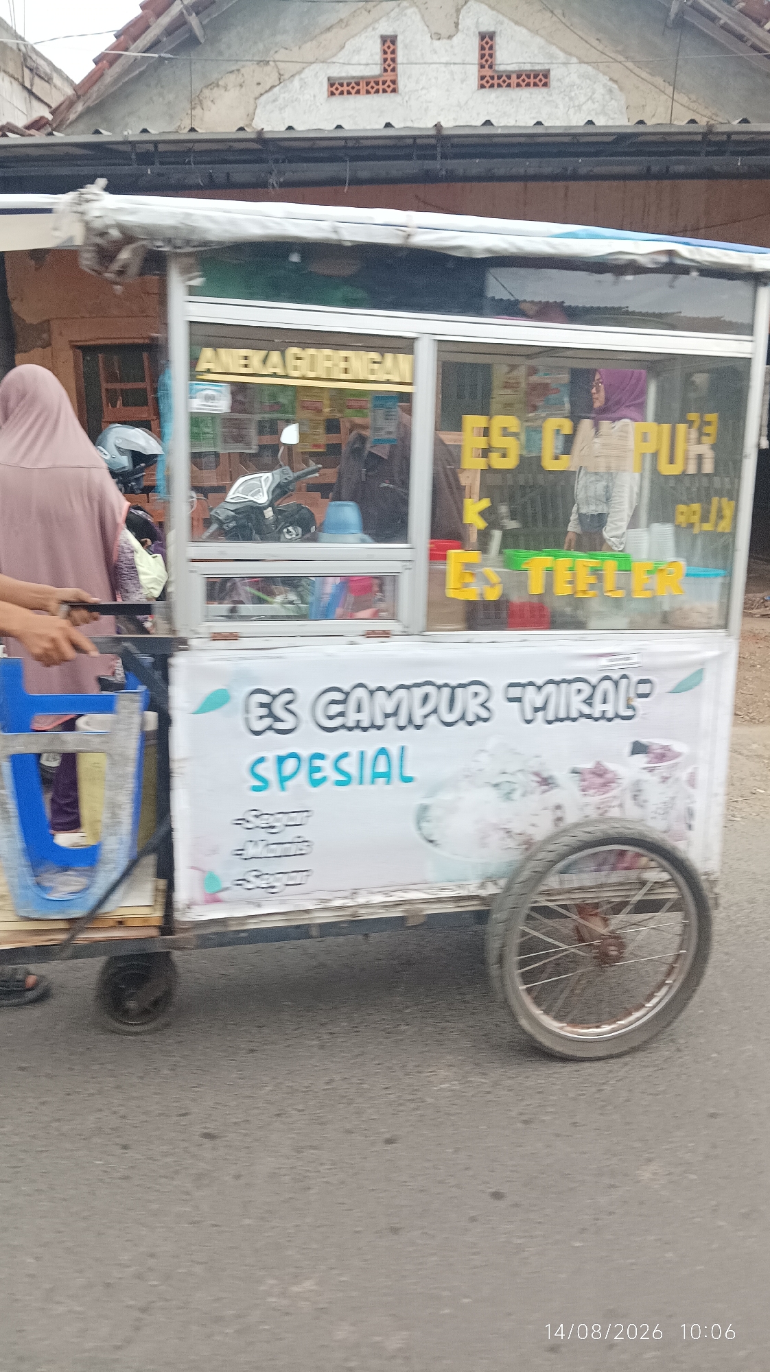 The image shows a food cart selling "Es Campur 'Miral'" (Mixed Ice "Miral") and "Aneka Gorengan" (Assorted Fried Foods) on a street in Tasikmalaya, Indonesia. The cart is white with blue accents and has glass panels displaying the food items. A woman in a brown hijab is standing behind the cart. The cart has a wheel on the right side. In the background, there is a building with a tiled roof and a motorcycle parked behind the cart. The street is paved. A timestamp at the bottom right corner reads "14/08/2026 10:06".