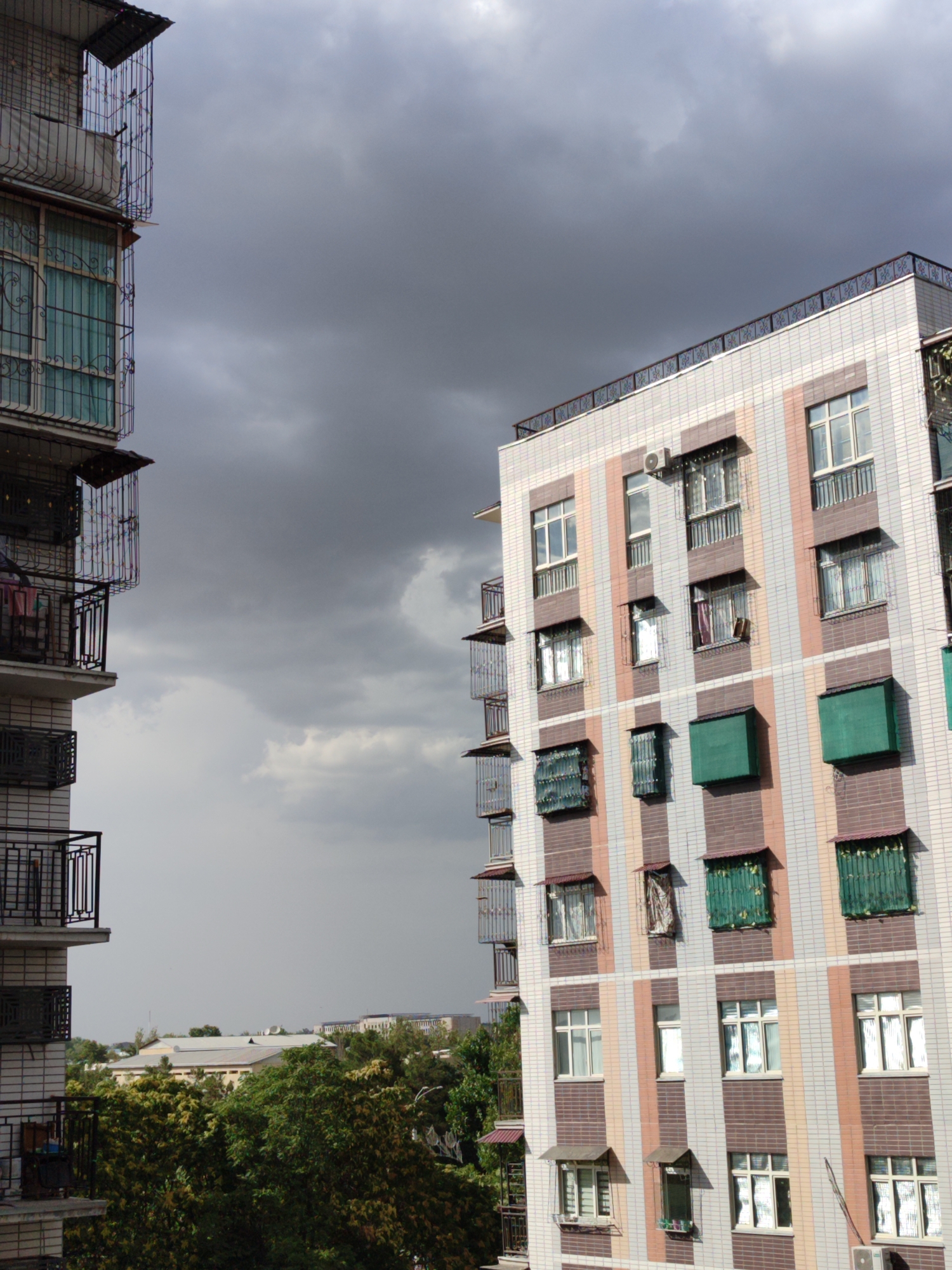 This image shows an exterior view of two apartment buildings in Minglola mahalla, Uzbekistan, under a cloudy sky. The building on the right is a modern, multi-story structure with a facade of pink, white, and brown tiles arranged in vertical stripes. It features numerous windows, many with green awnings or metal coverings. Balconies with black railings are visible, some with laundry hanging. An air conditioning unit is mounted on the building's exterior.
The building on the left appears older and is characterized by its light-colored brickwork and extensive use of decorative metalwork on its balconies and window guards. Laundry is also visible hanging from some of these balconies.
In the background, lower buildings and lush green trees are visible, suggesting a residential area within the city. The sky is overcast, indicating potentially poor weather. No people are visible in the image.