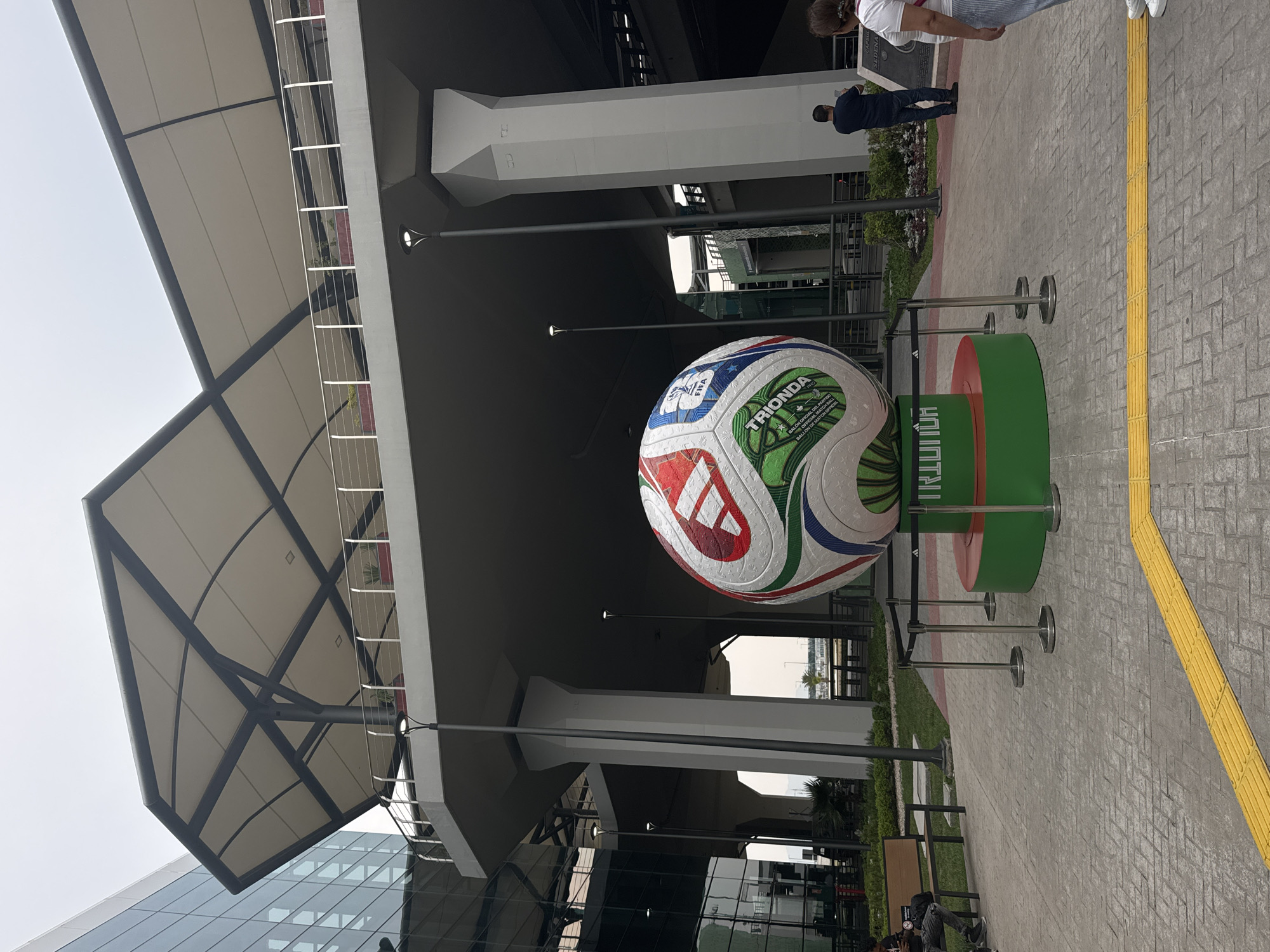 The image depicts a public space in Santa Lucía, Mexico. A large, oversized soccer ball, branded "TRIONDA" and "FIFA," rests on a green and red circular stand, positioned near a pedestrian walkway. In the background, a building with a modern architectural design features a large awning supported by white pillars. On the paved walkway, a yellow tactile paving strip for the visually impaired is visible. Several individuals are present, including a person in blue walking away from the camera, and two others partially visible near the upper right corner. Glass windows, greenery, and lamp posts further define the environment.