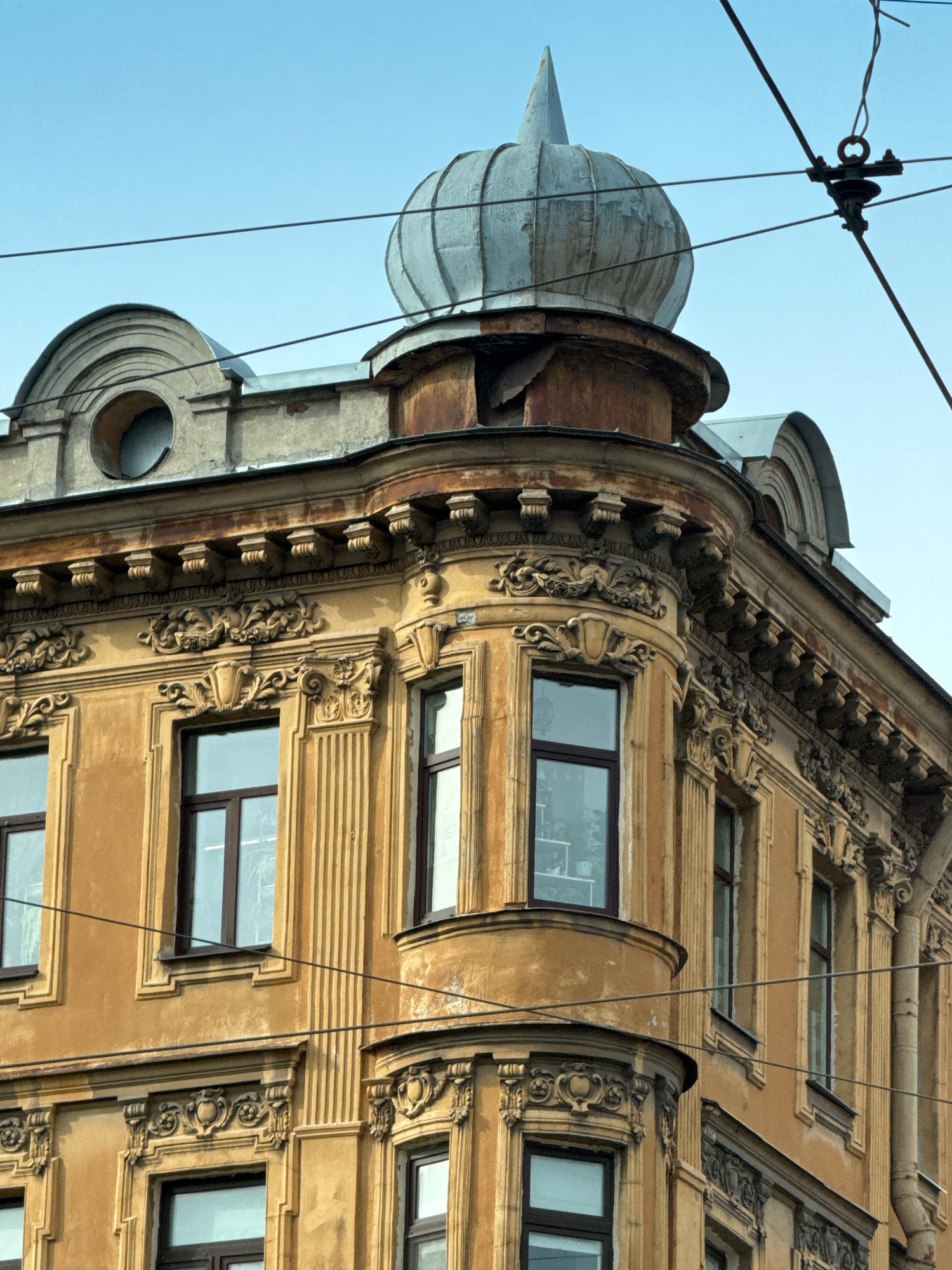 The image shows the upper corner of an ornate, yellow-ochre building with classical architectural details in Saint Petersburg, Russia. The building features decorative moldings, pilasters, and several windows with dark frames. A rounded bay window is visible on the corner. Above the main facade, a domed turret with a pointed finial crowns the structure, constructed from weathered metal. The sky is a clear, pale blue, and overhead, thick electrical wires crisscross the frame, suggesting an urban environment. The building appears aged, with some signs of wear and tear on the facade and the metal dome.