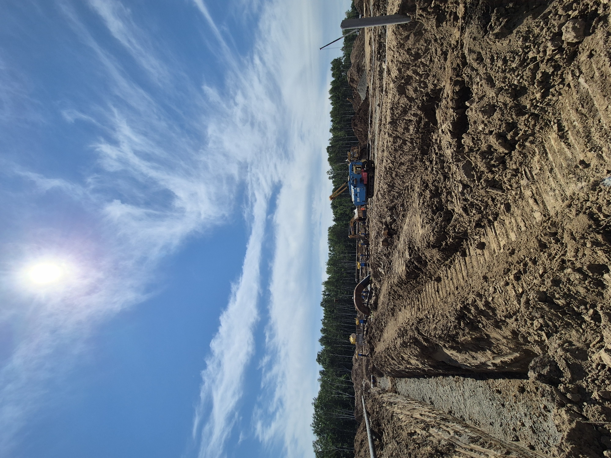 The image shows an outdoor construction site in Taezhnoe, Russia. The sun is high in the sky, creating a bright glare. The sky is a vibrant blue with wispy white clouds. The foreground of the image is dominated by a large trench filled with gravel. The ground is composed of dark brown soil, with visible tire tracks from heavy machinery. In the middle ground, there are various construction vehicles, including a blue and red tracked vehicle with the text "2450 CH-24" visible, an excavator, and other machinery. Large pipes are also visible, including a large curved pipe and several smaller ones. A line of trees forms the background of the scene, suggesting the site is near a forest. A tall, grey pole stands on the right side of the image. The overall impression is one of active construction and land development.