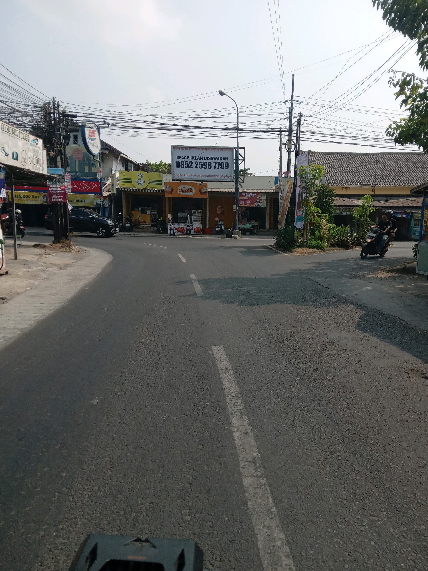 The image depicts a street scene in Banguntapan, Indonesia. A paved road curves into the distance, with a white dashed line marking the center. On the left side of the road, a sidewalk runs alongside a row of businesses, including a car wash and a shop with a sign reading "MOCI JAYA SNACK". A black car is parked on the left side of the road, and several motorcycles are visible parked near the businesses. On the right side of the road, there are more businesses, including one with a yellow awning and a sign for "BAKER OLI". A billboard advertising "SPACE IKLAN DISEWAKAN" (Space for Rent) with a phone number is prominently displayed. A lamppost stands near the billboard. Further down the road on the right, two individuals are riding a motorcycle. Trees and power lines are visible throughout the scene, with a dense network of wires overhead. The sky is clear and bright, suggesting a daytime setting.