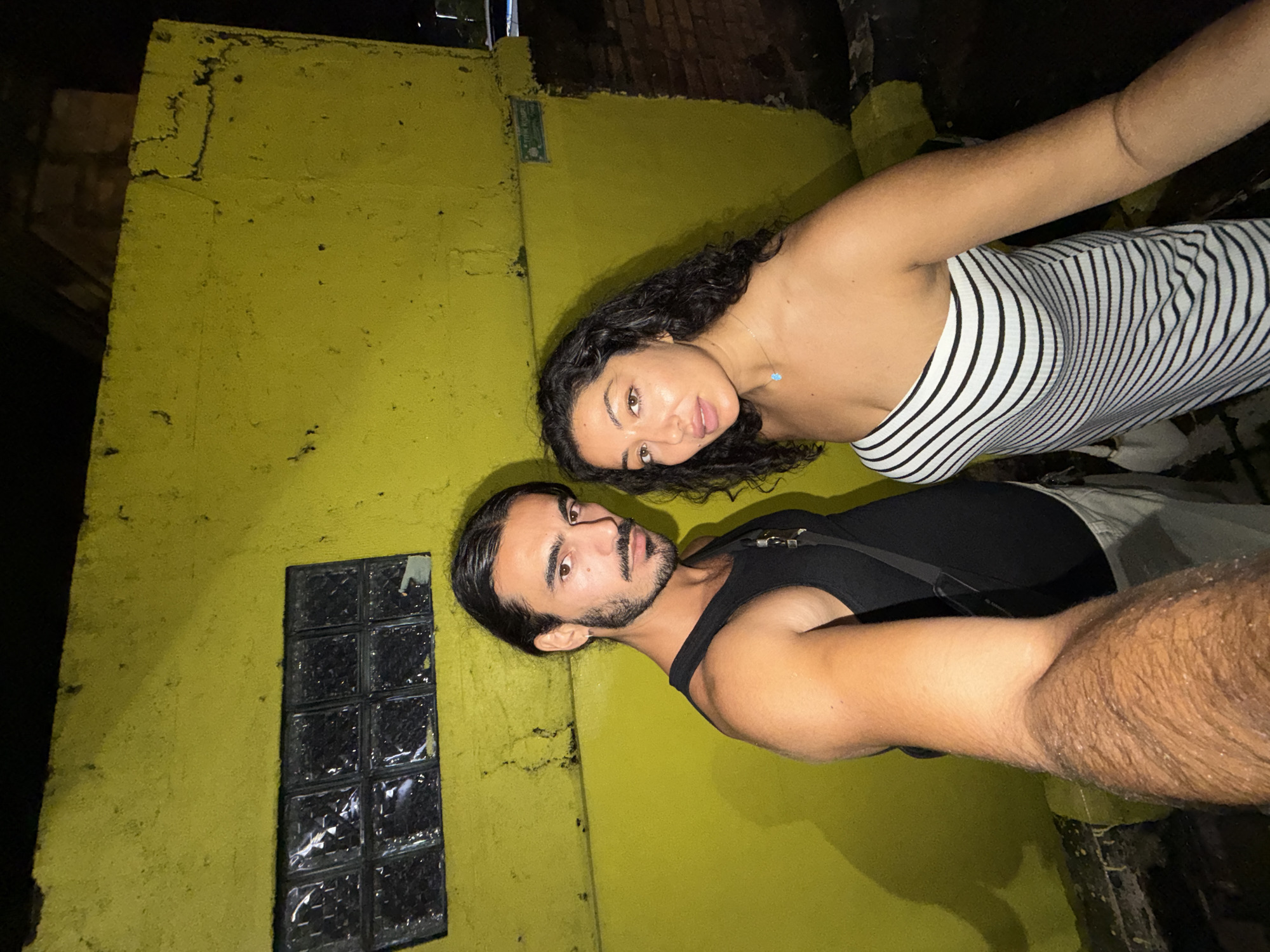 Two individuals, a woman and a man, pose for a close-up photo against a bright, textured yellow wall, likely at night in North Kuta, Indonesia. The image is tilted, suggesting it was taken as a selfie.
The woman, on the right, has dark, curly hair and brown eyes. She wears a black and white striped strapless top and a delicate silver necklace with a small, light blue pendant. Her expression is neutral as she looks towards the camera.
The man, on the left and slightly below the woman, has dark hair pulled back, a beard, and a mustache. He wears a black tank top or sleeveless shirt, revealing muscular arms. He also looks directly at the camera with a neutral to slightly serious expression.
The background consists primarily of a vivid yellow wall, which shows signs of wear, including dark stains and irregular textures, especially at its edges. Embedded in the wall to the left is a small, rectangular window composed of multiple glass blocks. Above the woman's head, in the shadows of an overhang, faint dark brickwork is visible, hinting at an older structure. There's a small, rectangular, white plaque-like object behind the woman's head on the wall, but any text on it is illegible. The overall lighting is dim and artificial, casting shadows and highlighting the subjects against the brightly painted wall.